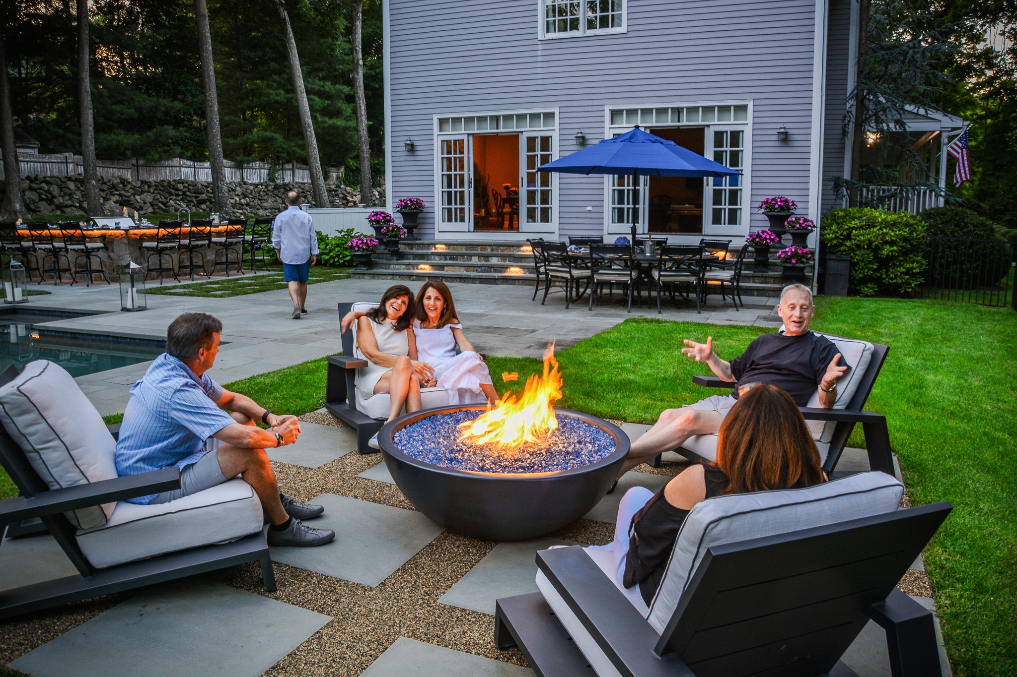 A group of people is relaxing around a fire pit in a backyard with a pool and outdoor dining area in the background. A person is walking away.