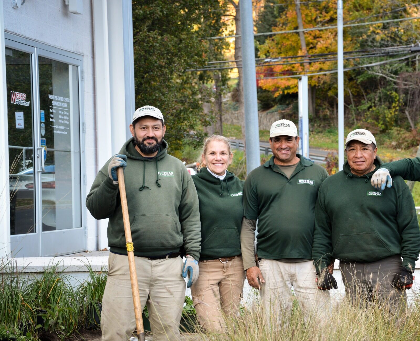 Five people in green uniforms stand together outdoors, smiling, with landscaping tools, by a garden bed next to a building.