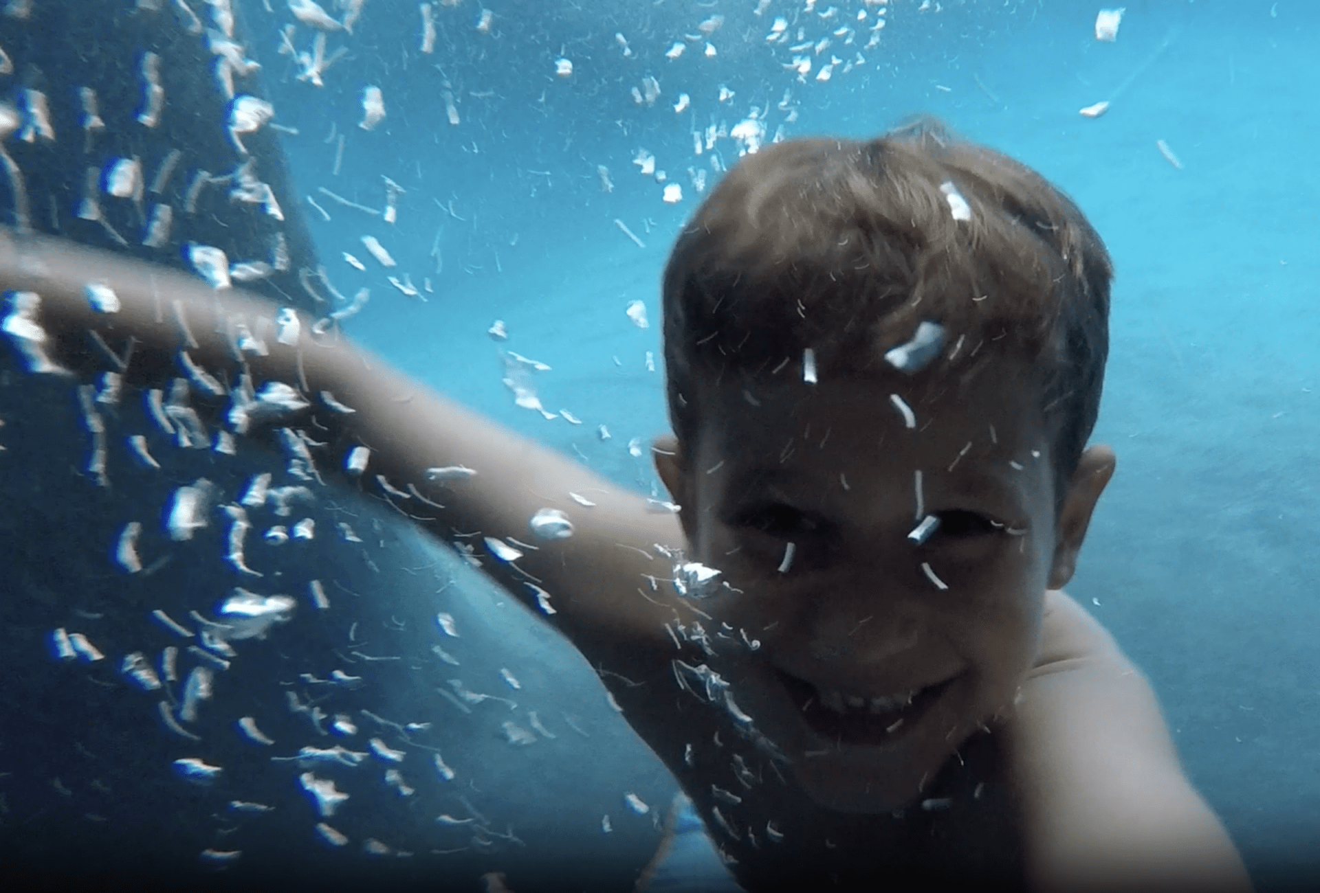 A smiling child is submerged in clear blue water, surrounded by bubbles, with a joyous expression visible through the water's distortion.