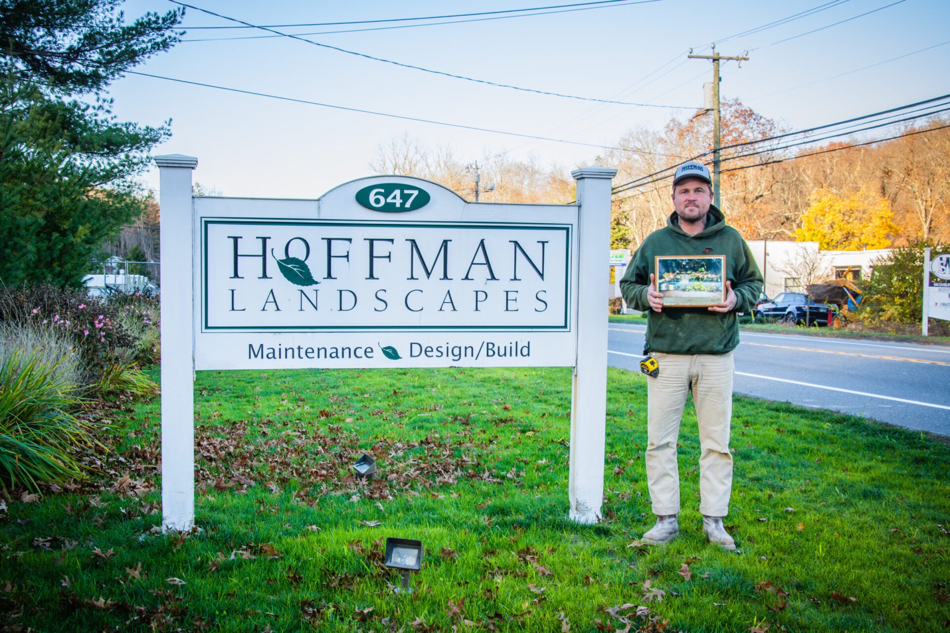 A person stands by a sign reading "HOFFMAN LANDSCAPES" holding a plaque and a measuring tape, on a roadside with greenery and a clear sky.