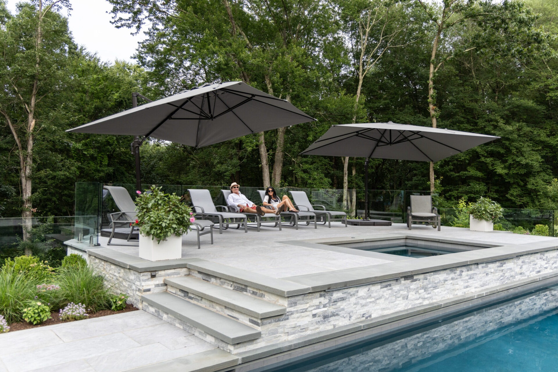 This image shows a luxurious poolside area with two persons lounging on chairs under umbrellas, surrounded by lush trees and clear skies.