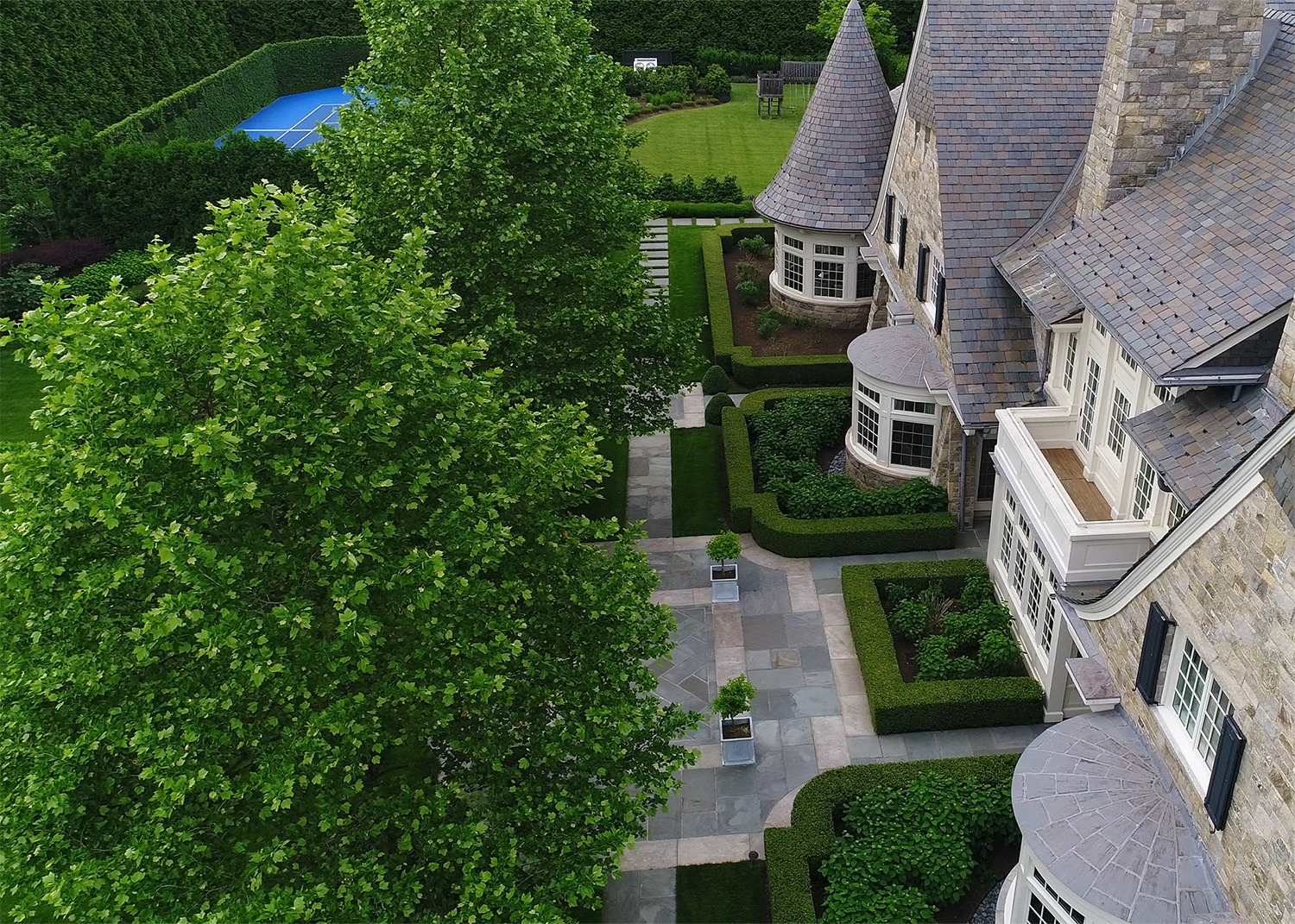 An aerial view of a luxurious estate with manicured gardens, hedges, stone pathways, a swimming pool, and architectural details on the roof.