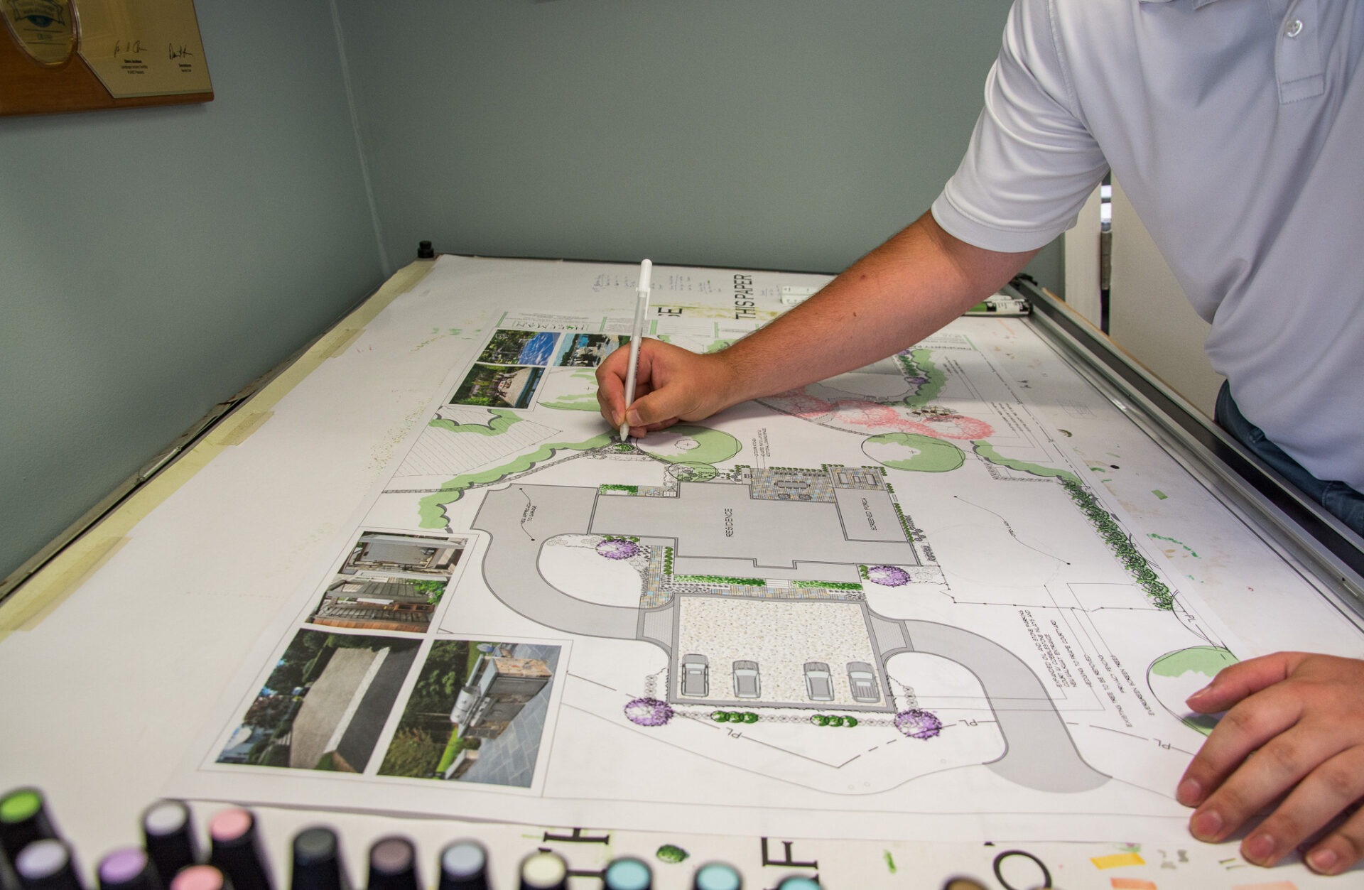 A person is drawing on a landscape design plan with colored markers. Various architectural details and reference photos are visible on the layout.