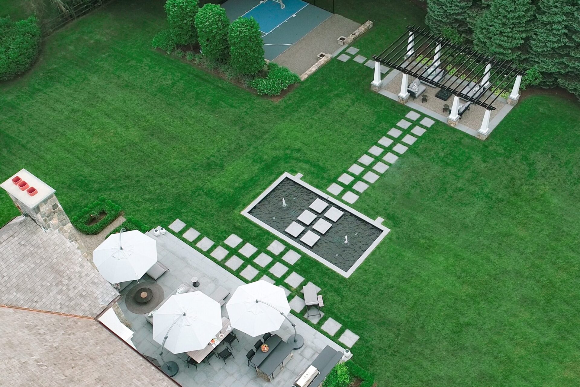 Aerial view of a landscaped backyard with a patio, umbrellas, seating areas, a basketball court, ornamental garden, lawn paths, and a decorative pergola.