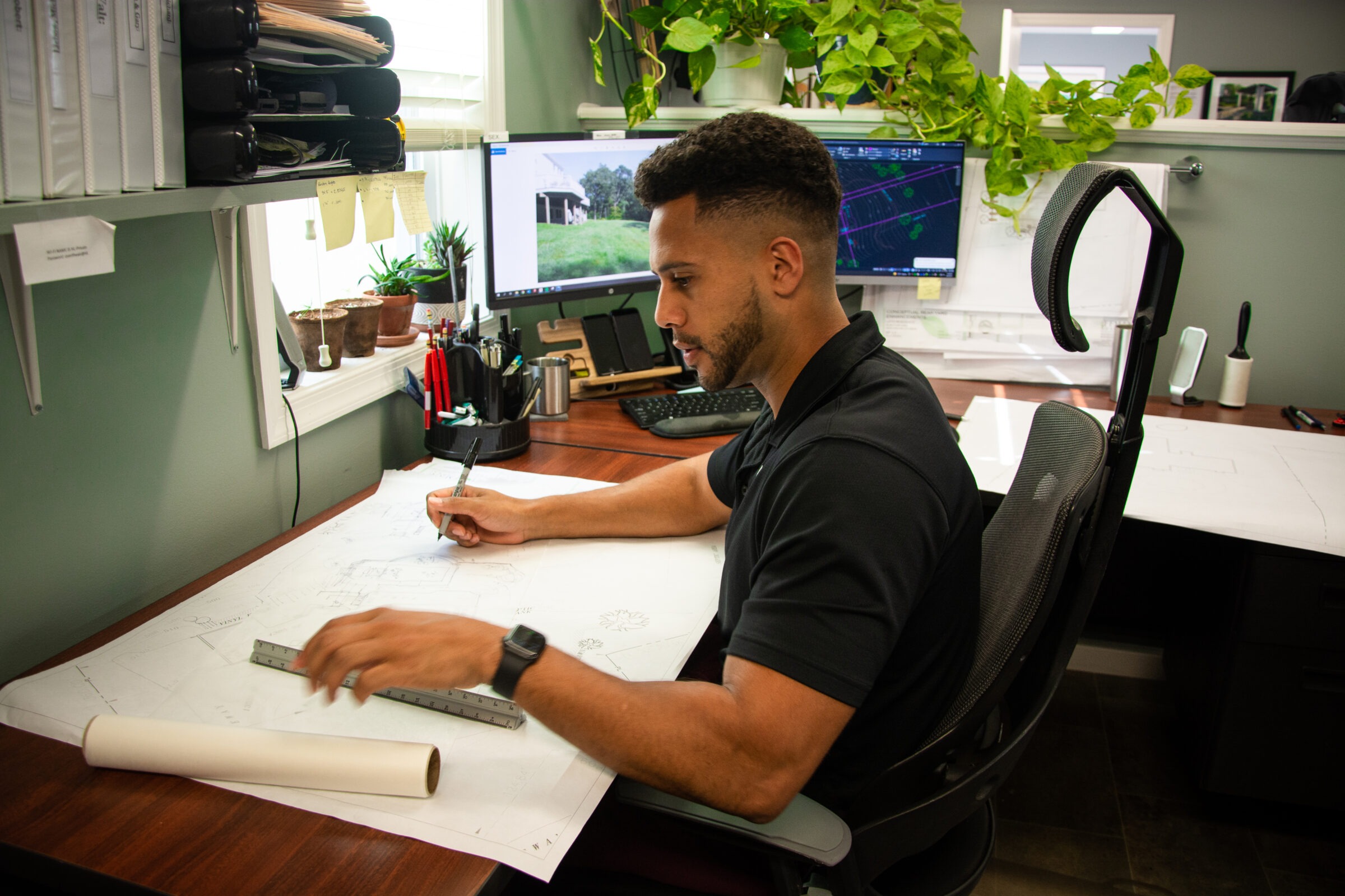 A person is working at a desk with architectural plans, a ruler, and pens. Computer screens and office supplies are visible in a well-lit room.