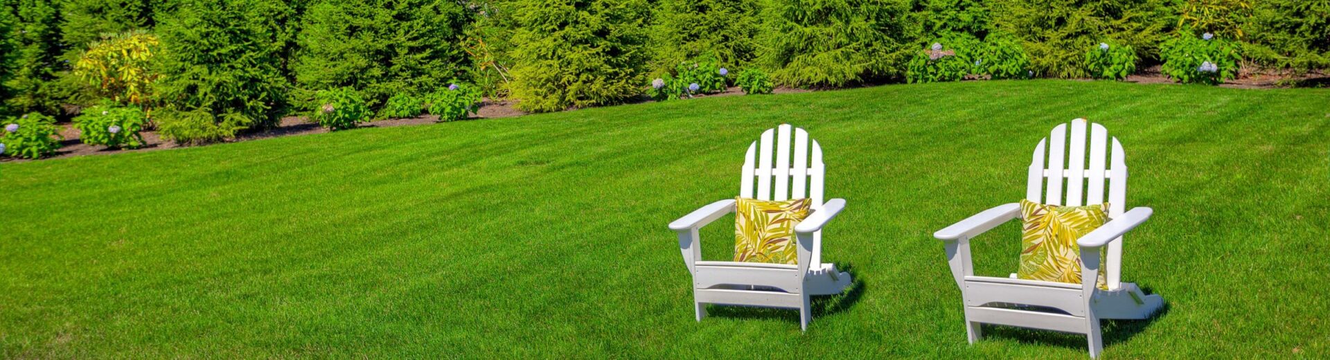 Two white Adirondack chairs with yellow patterned cushions are set on lush green grass, facing a landscaped garden with dense shrubs and flowers.