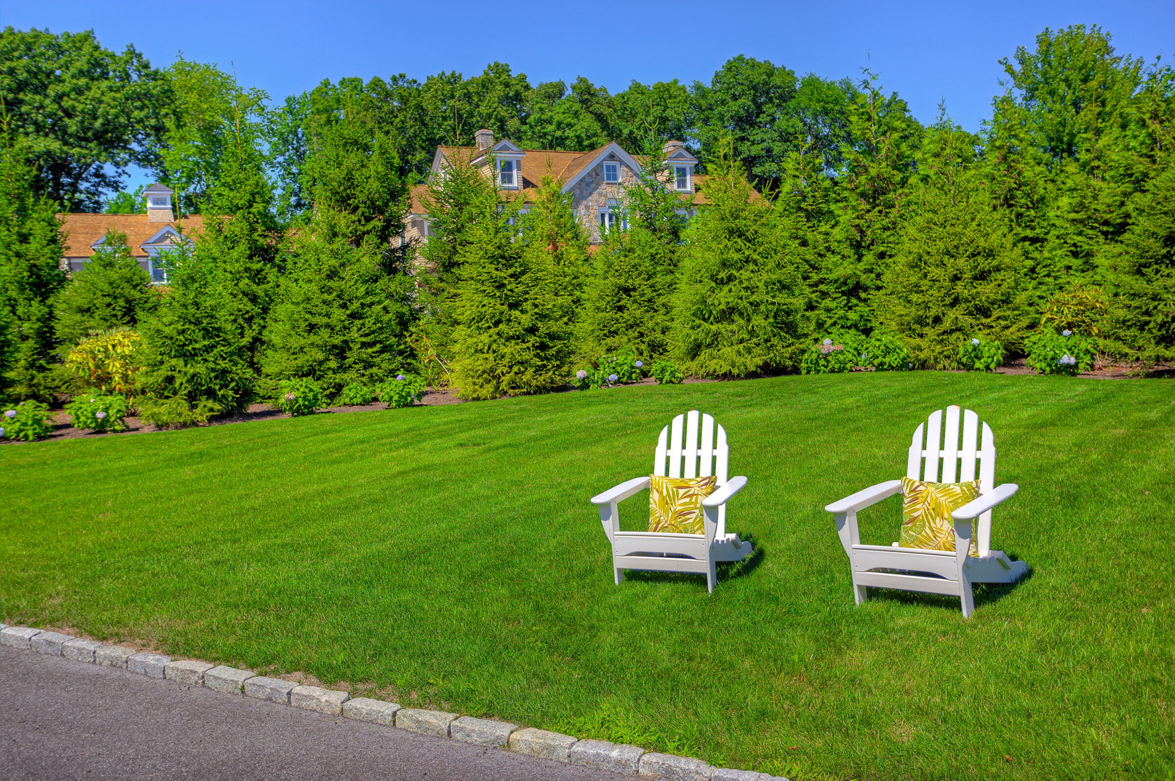 Two white Adirondack chairs with cushions sit on lush green grass, overlooking a large house with dormer windows nestled among tall green trees.