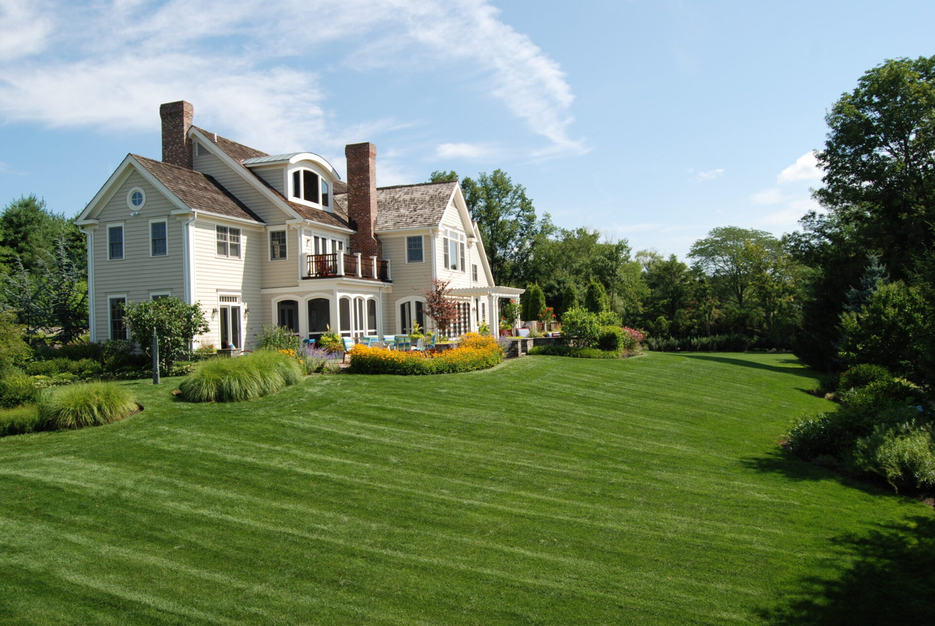 A large, elegant two-story house with a well-manicured lawn, surrounded by lush landscaping, under a clear blue sky.