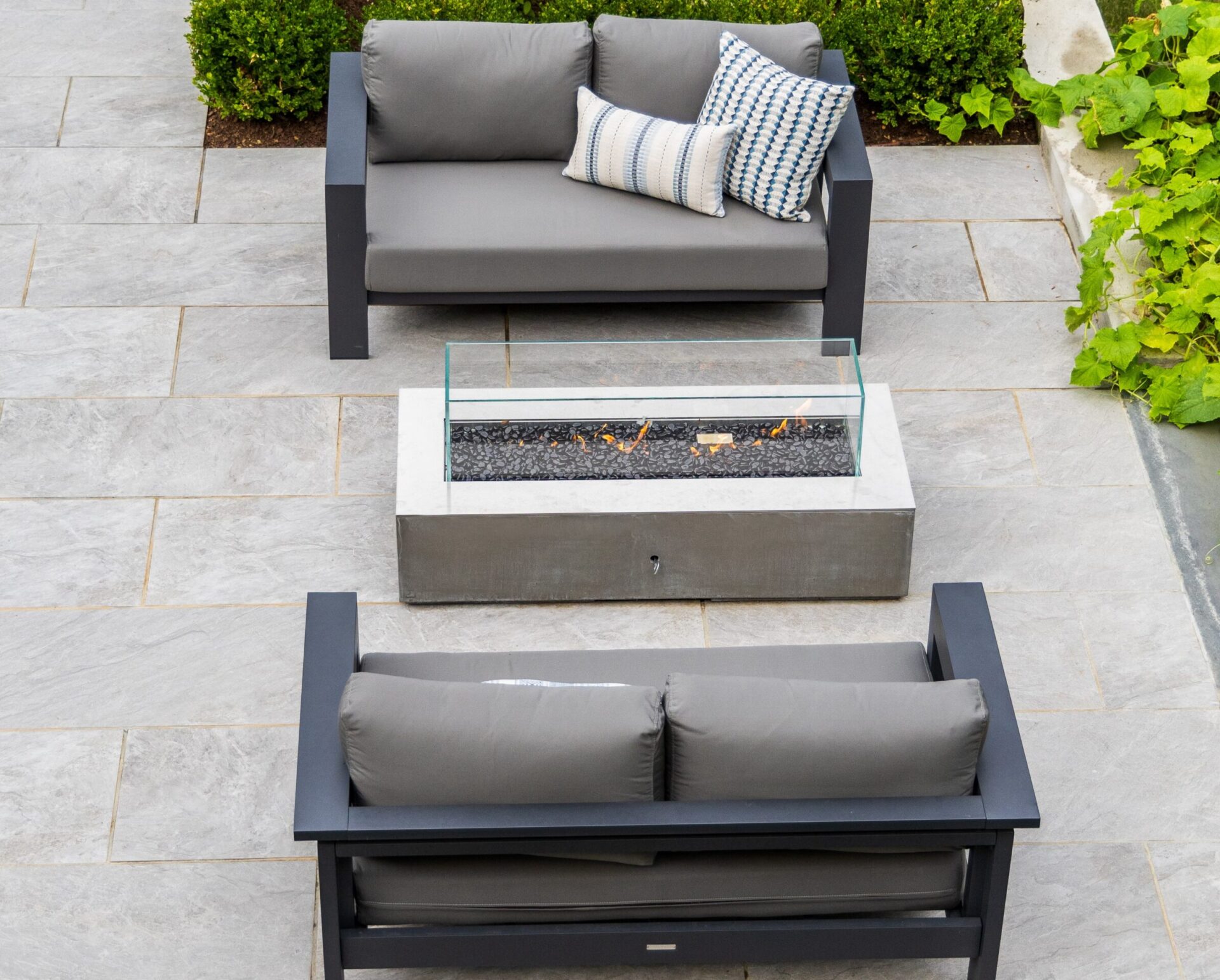 An outdoor setting featuring modern furniture with a fire pit table, two couches with cushions, on a tiled patio with surrounding green plants.