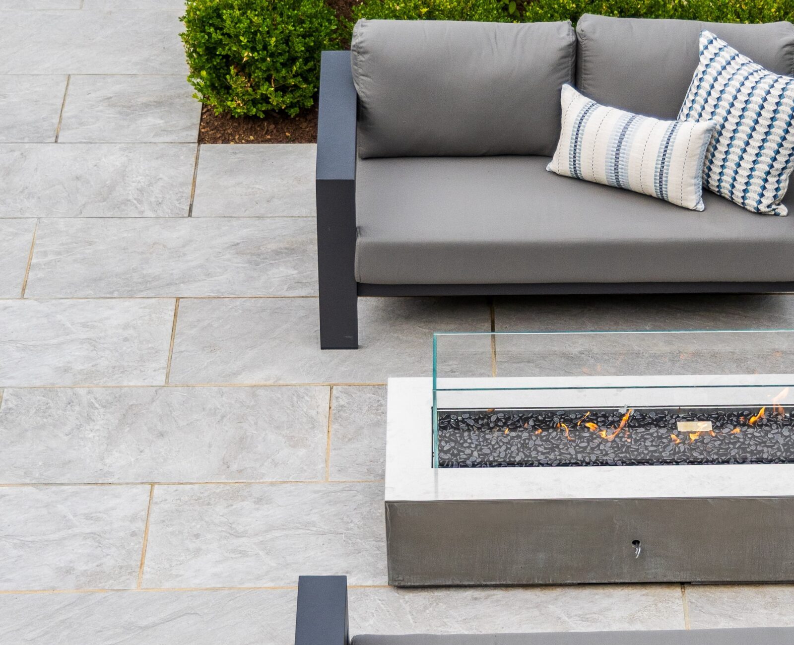 An outdoor setting featuring modern furniture with a fire pit table, two couches with cushions, on a tiled patio with surrounding green plants.