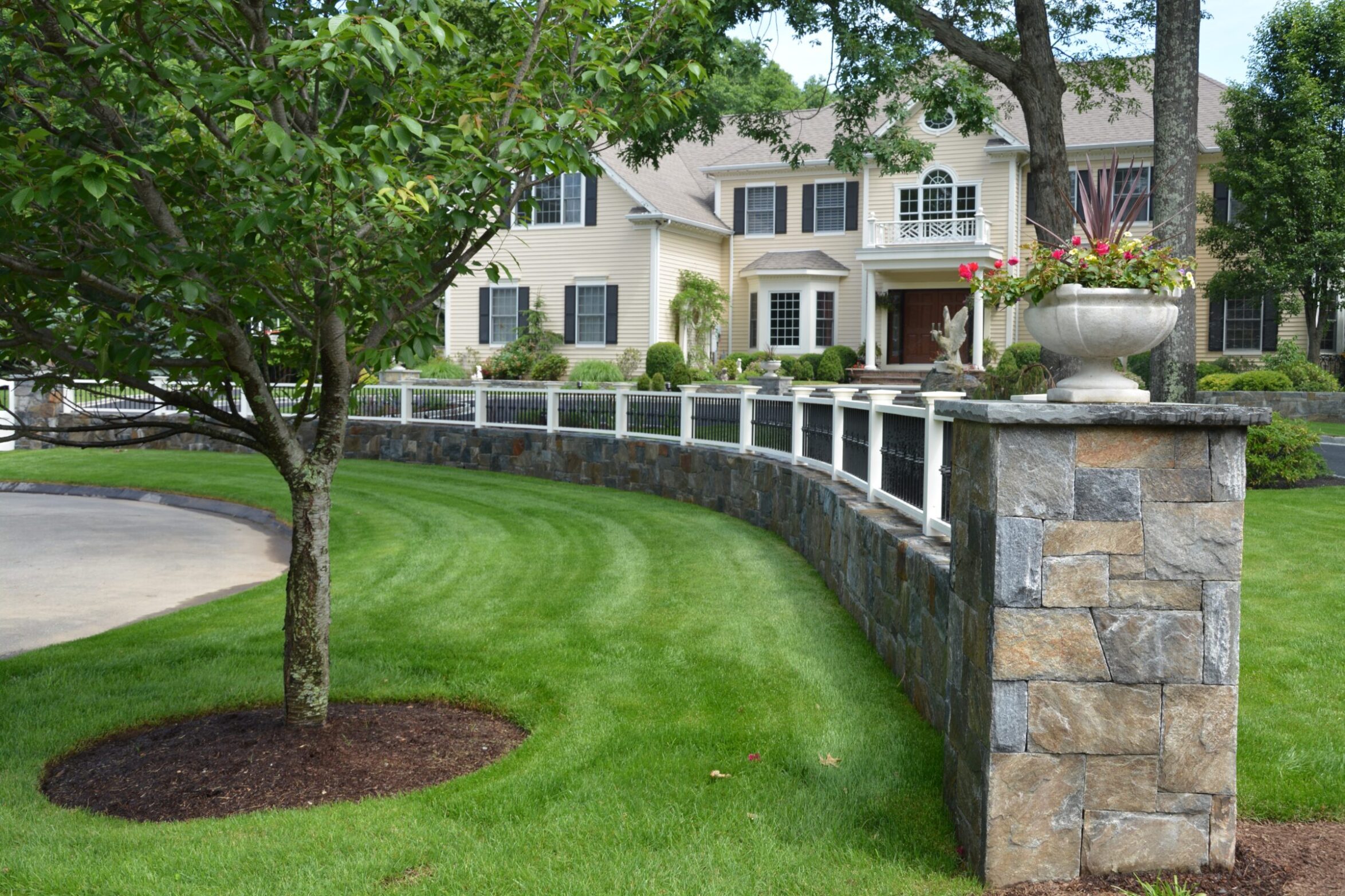 The image shows a manicured lawn with a stone and white railing fence, leading to an elegant, two-story house with a balcony and a planter.