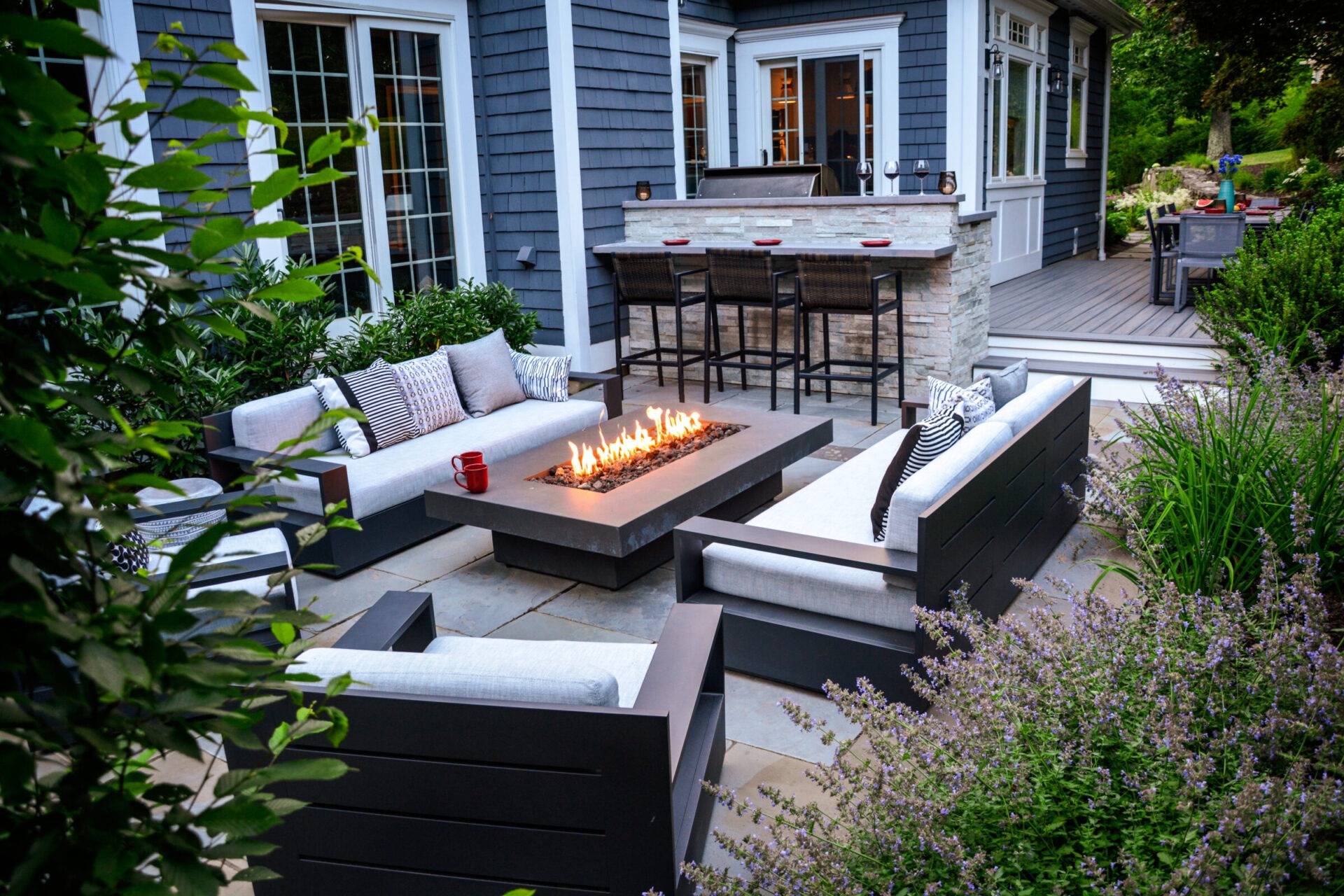 An outdoor living space with modern furniture, a fire pit, greenery, and a built-in grill bar against a backdrop of a blue-gray house.
