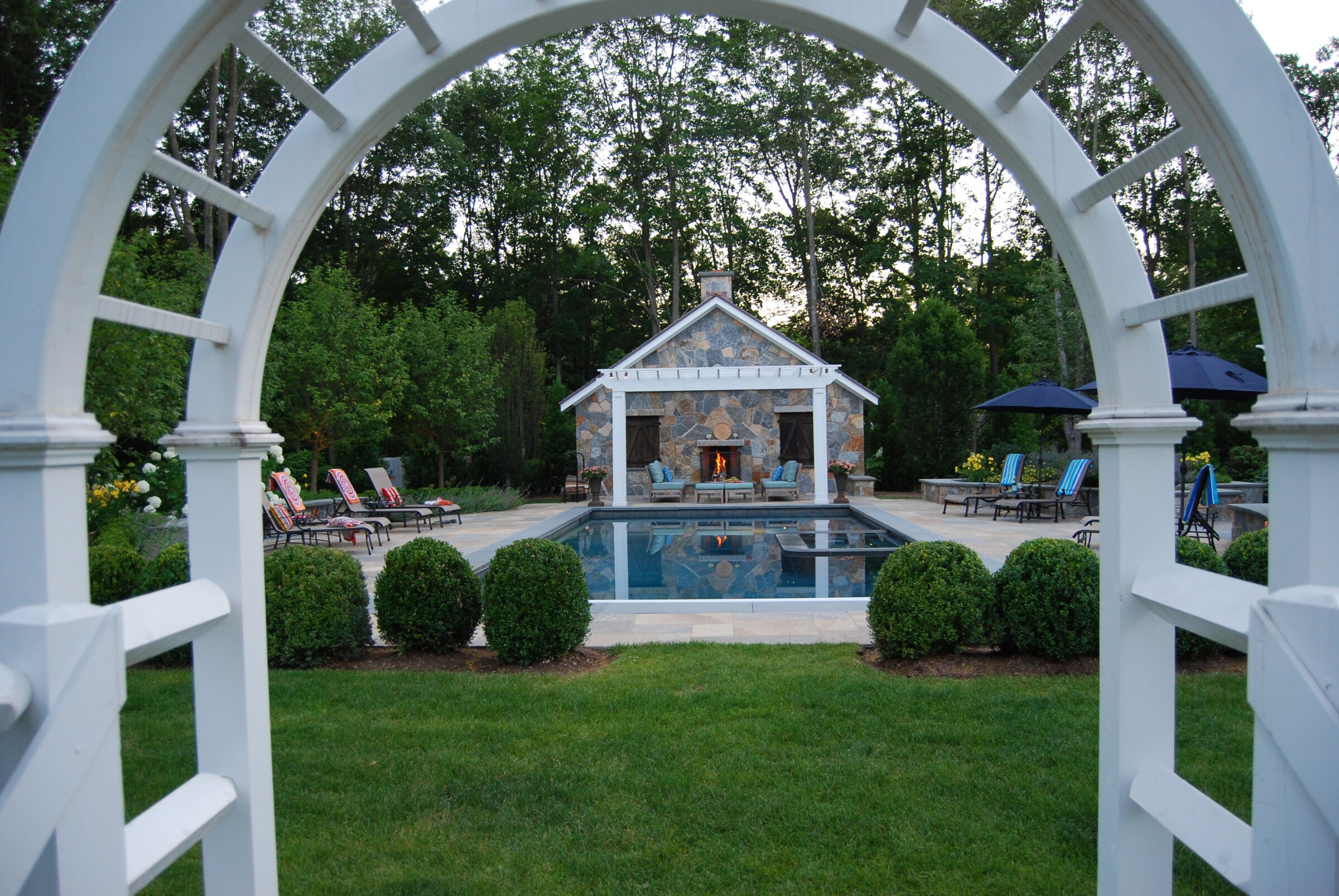 This image features an elegant backyard with a rectangular pool, a stone pool house with a fireplace, lounging chairs, umbrellas, and manicured greenery.