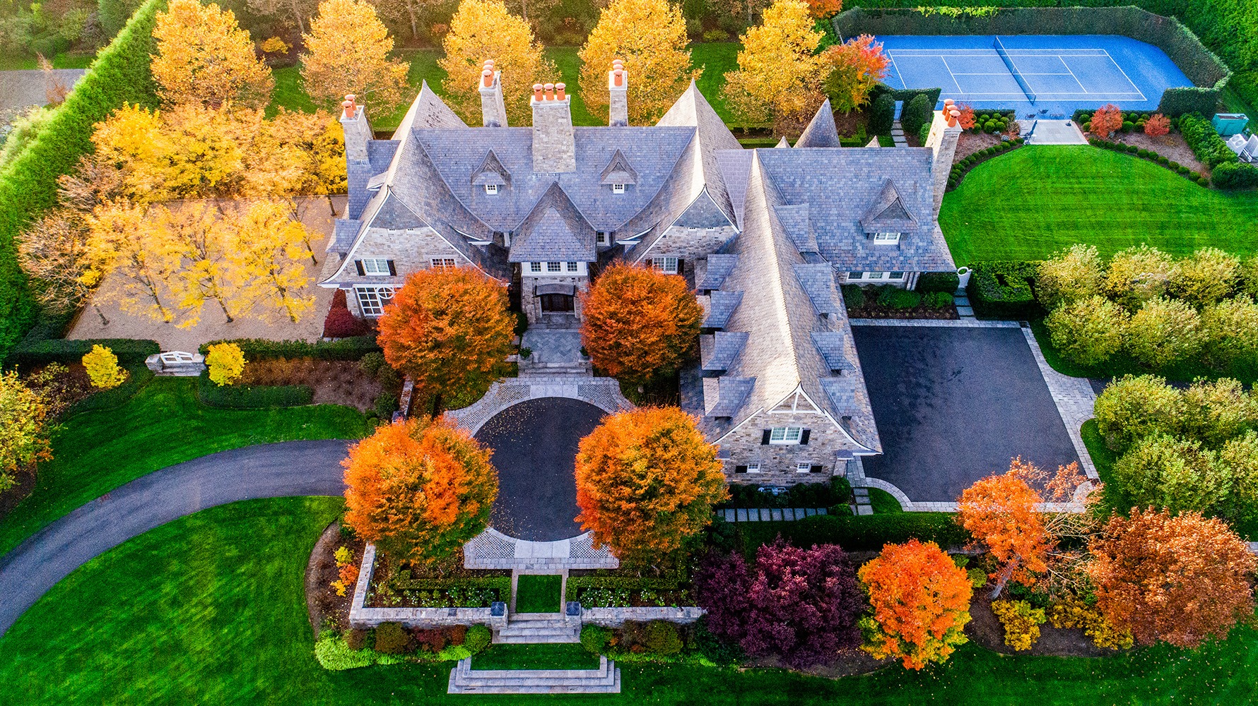 Aerial view of a large, elegant house with a slate roof, surrounded by colorful autumn trees, a manicured lawn, tennis court, and circular driveway.