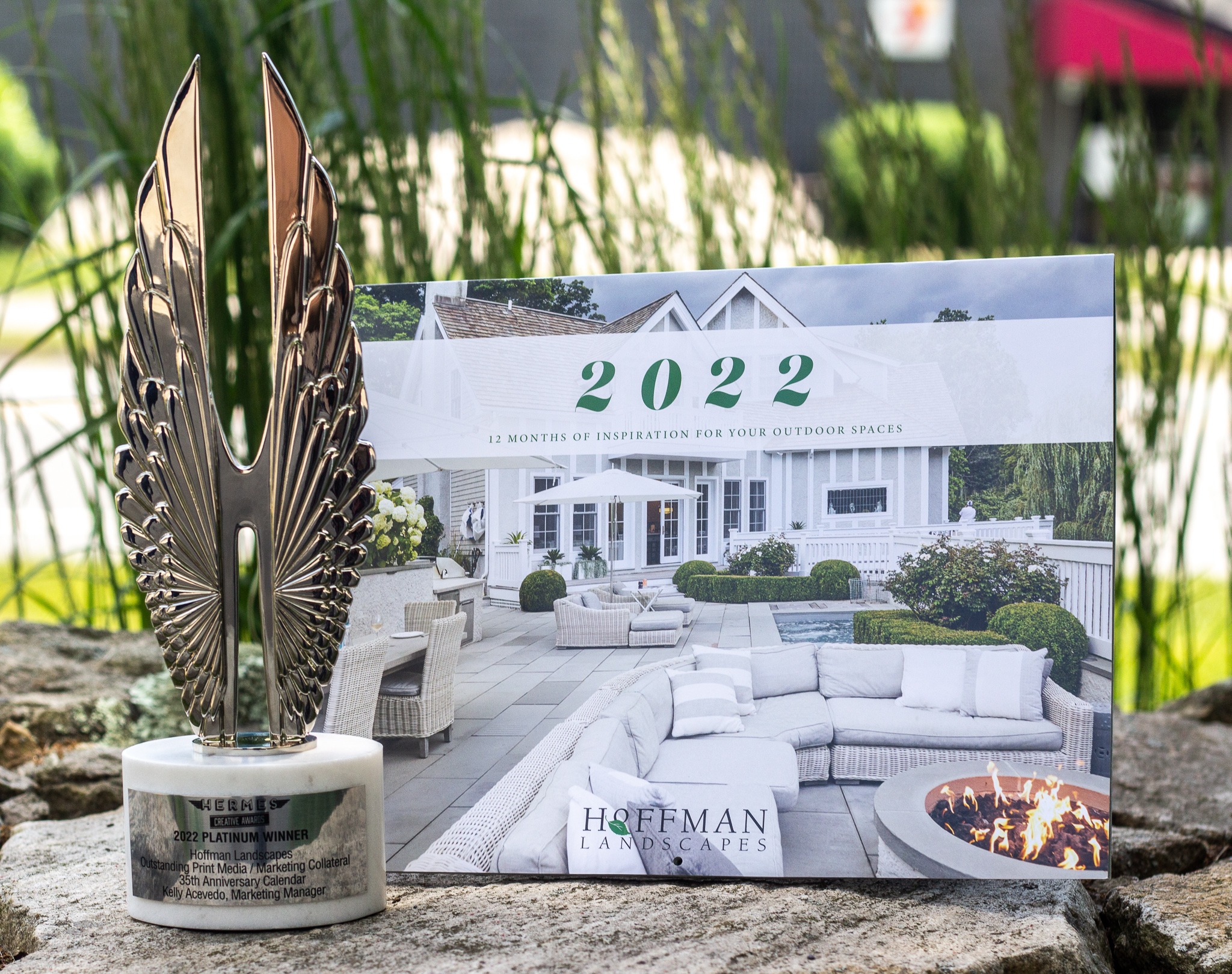 A trophy with feather-like details sits in front of a calendar showcasing outdoor living spaces, reflecting an award for marketing collateral. Greenery softly frames the background.