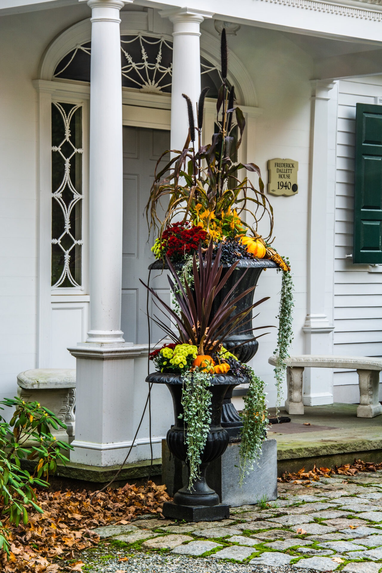 An ornate black urn on a pedestal filled with autumnal plants and vegetables, set against a white historic building with classical columns.