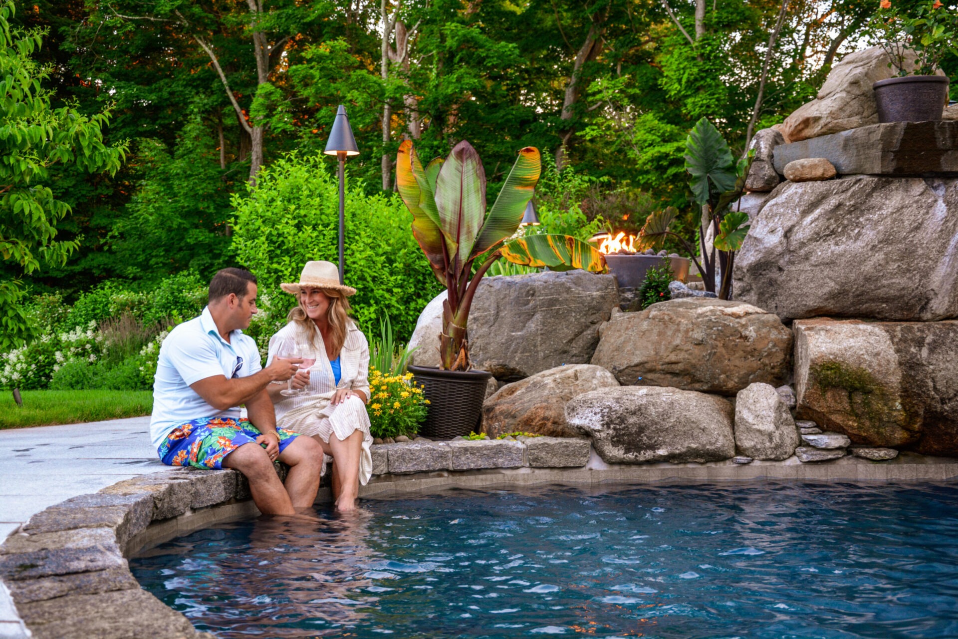 Two people are sitting by a pool with their feet in the water, enjoying drinks and smiling, surrounded by rocks, plants, and a fire feature.