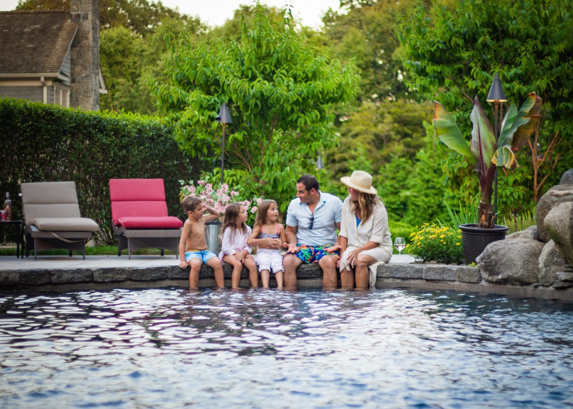 A group of two adults and three children sits by a poolside, chatting and enjoying a warm day in a lush garden with lounge chairs nearby.