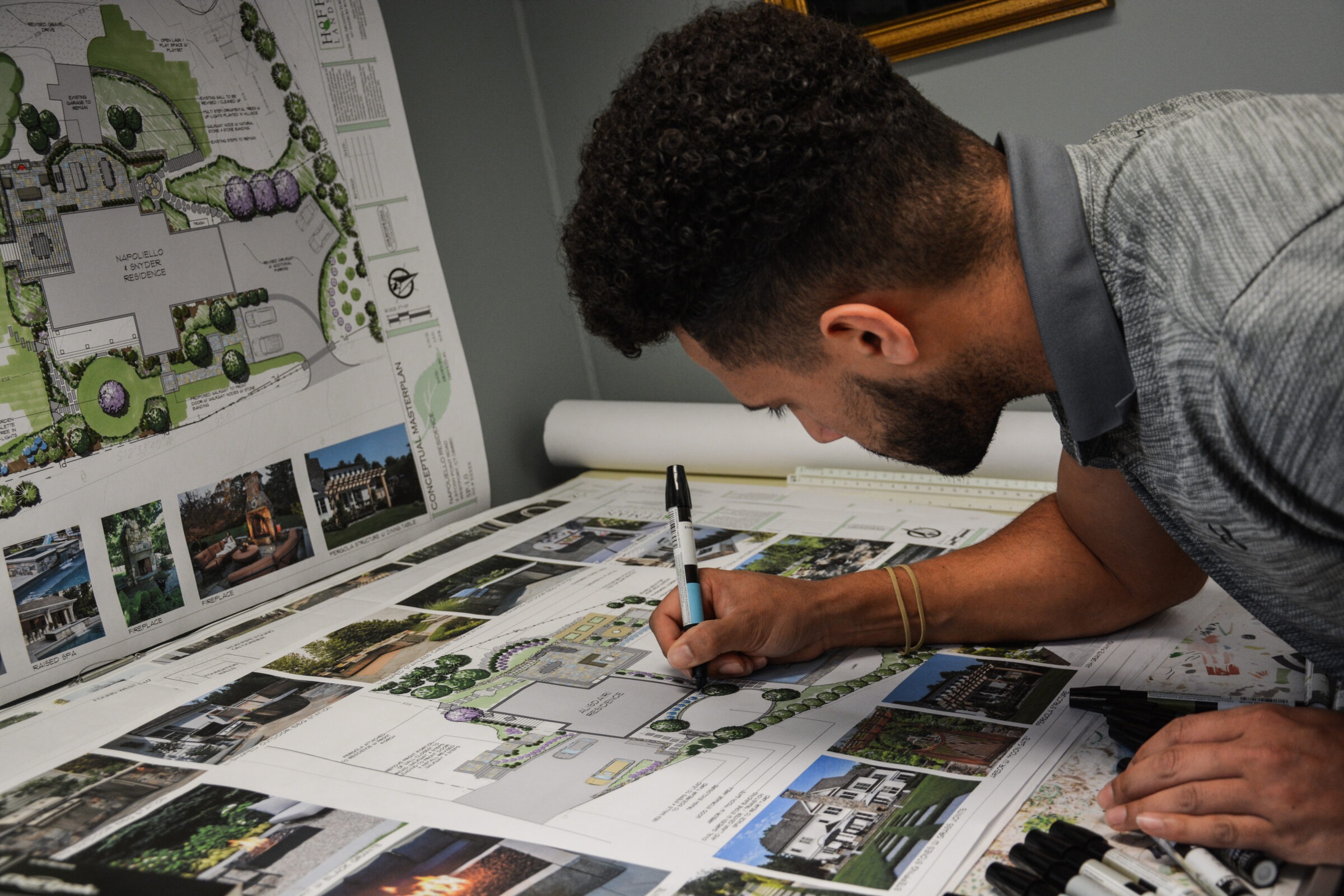 A person is intently working on architectural drawings and landscape plans spread across a desk, with markers scattered around.