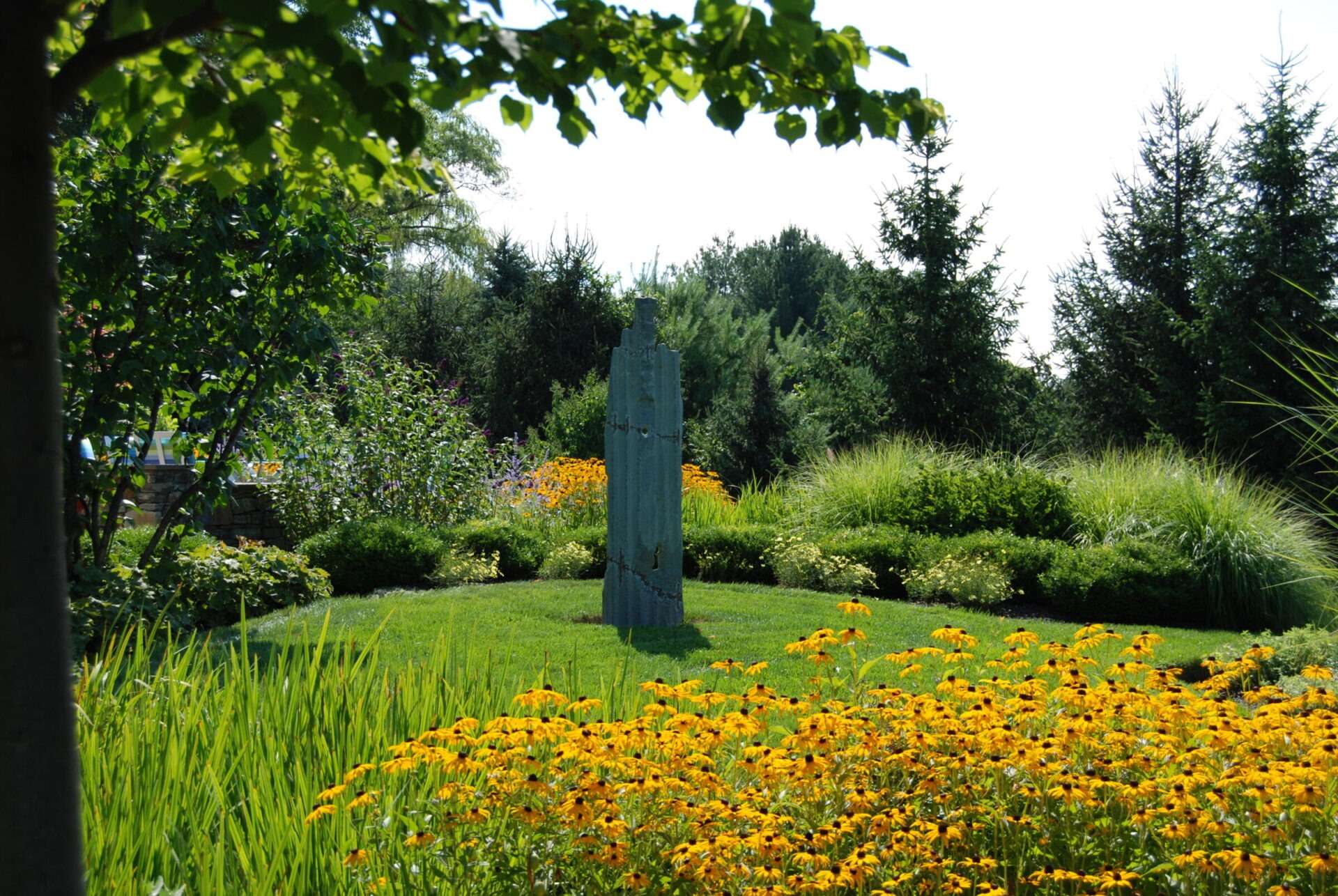 This image shows a vibrant garden with blooming yellow flowers, ornamental grass, green shrubs, and trees under a clear blue sky. An artistic sculpture stands centrally.