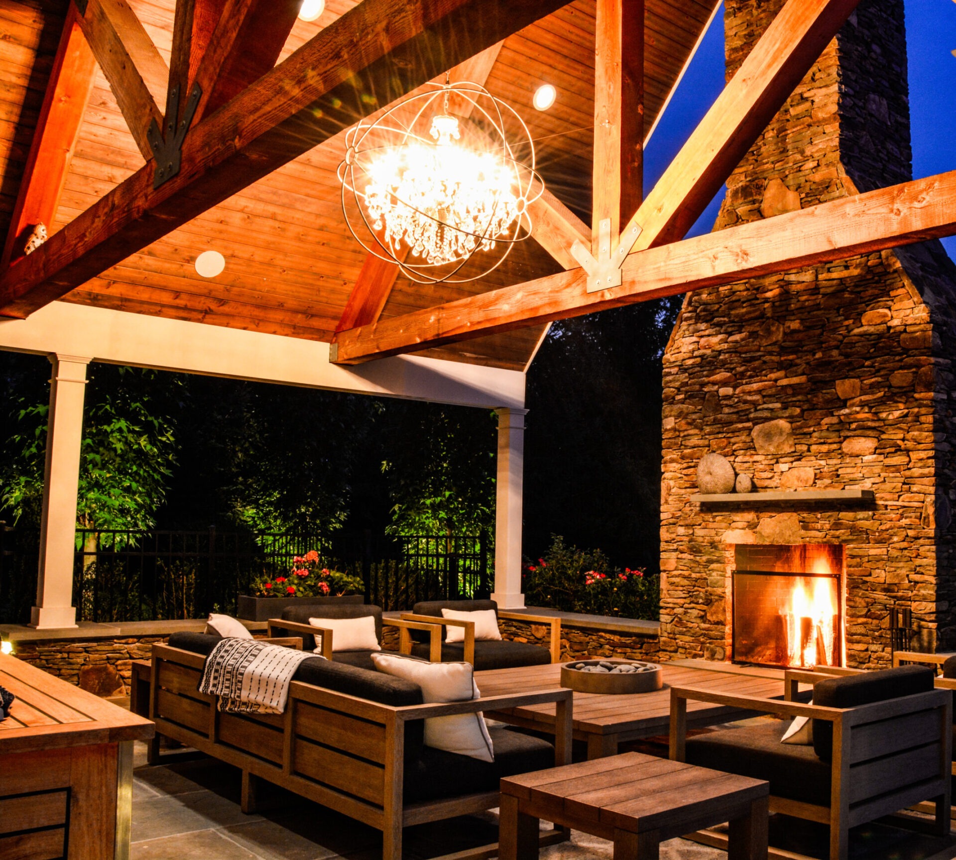 This image shows an outdoor covered patio at dusk with wooden furniture, a lit fireplace, a chandelier, and a warm, inviting ambiance.