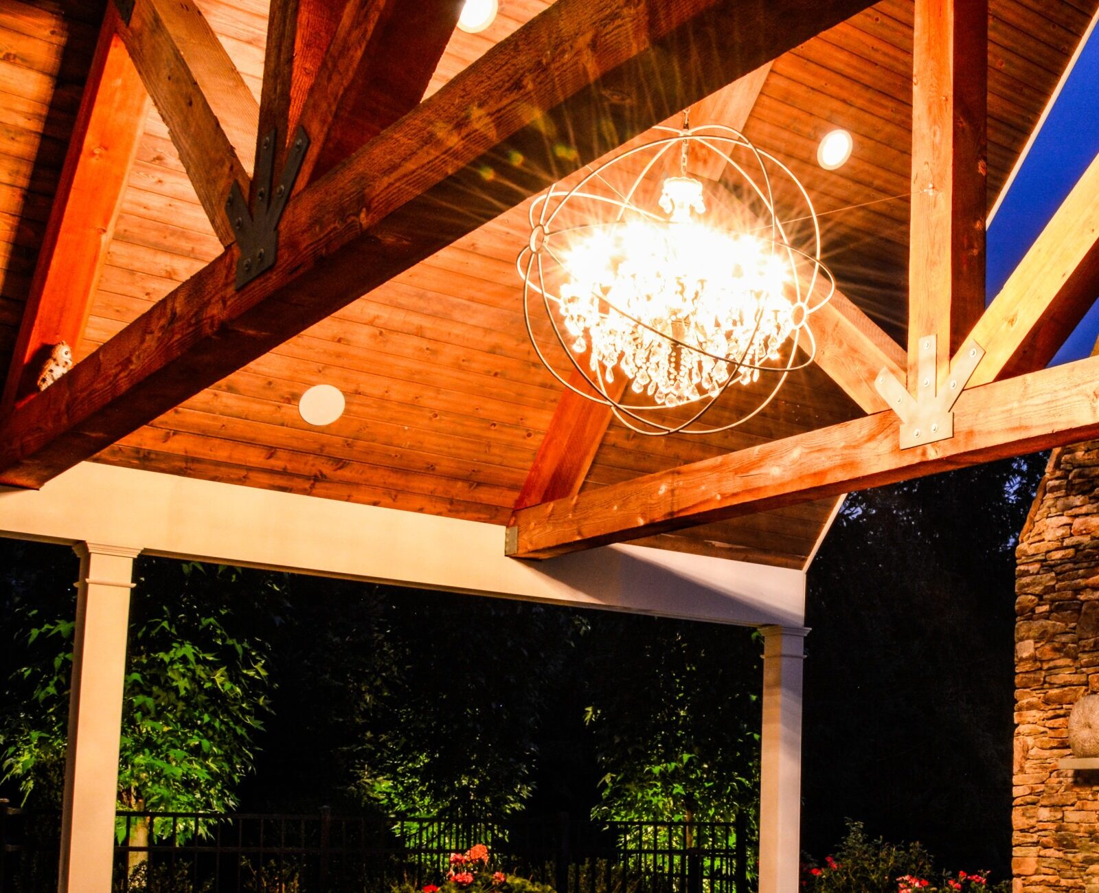 This image shows an outdoor covered patio at dusk with wooden furniture, a lit fireplace, a chandelier, and a warm, inviting ambiance.