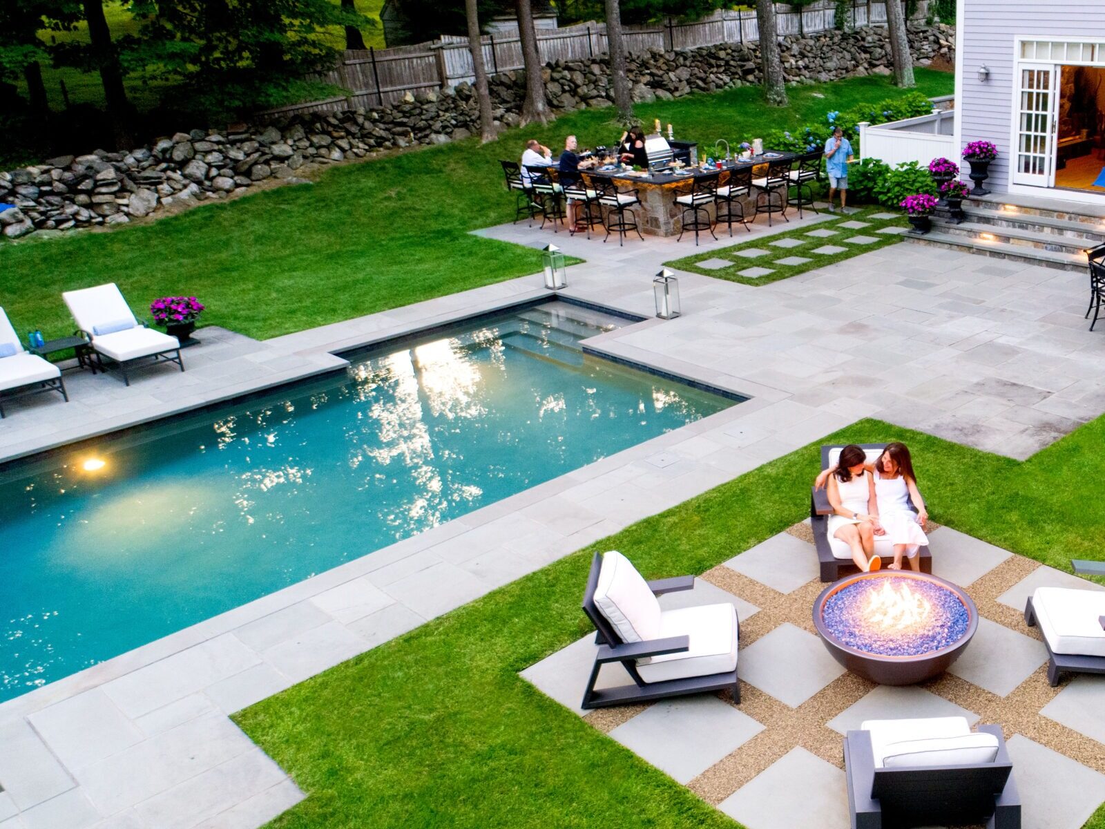 This image shows an aerial view of a backyard with a swimming pool, a seating area with a fire pit, loungers, and a dining area with people.