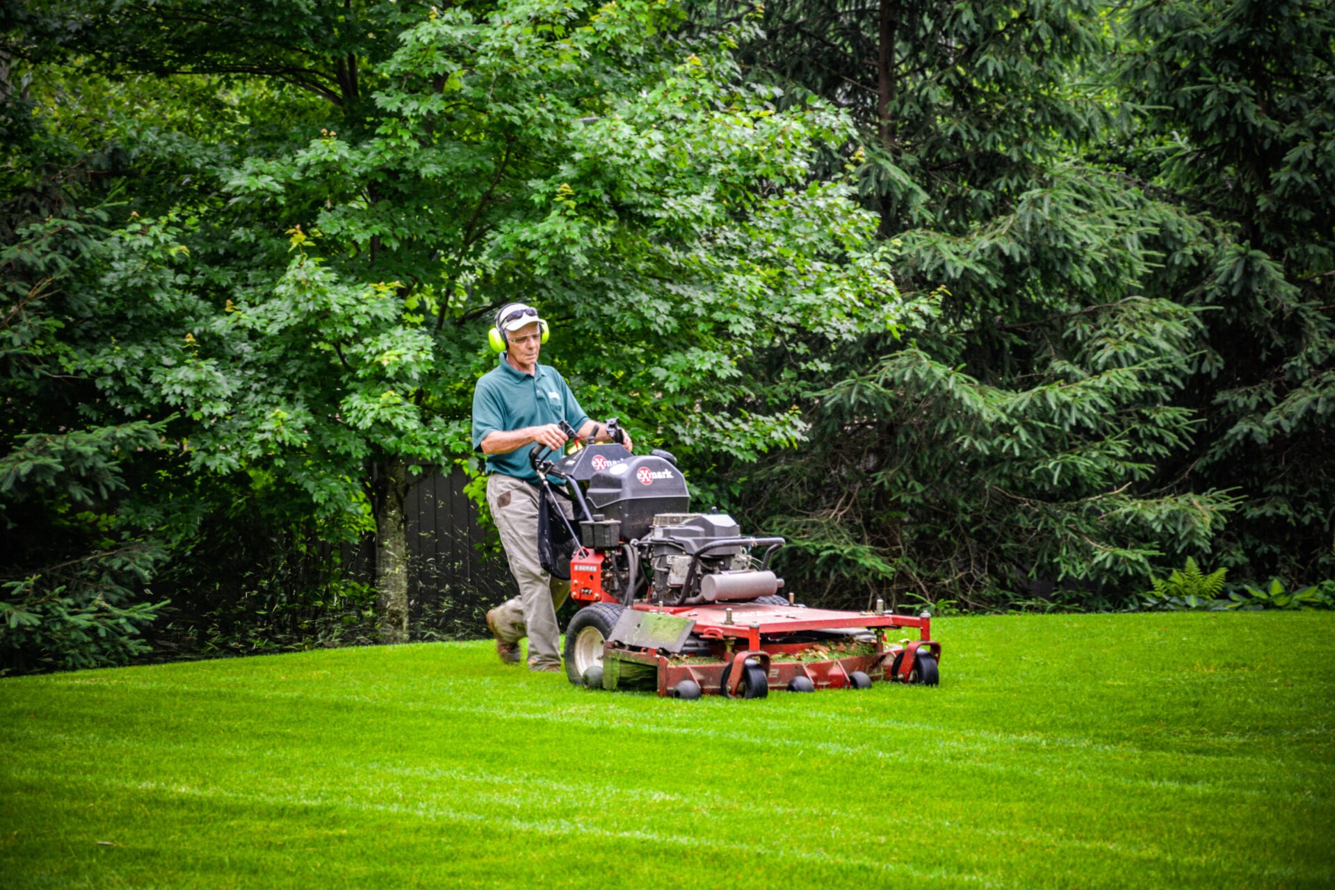 A person mows a lush green lawn using a red commercial riding lawn mower, with dense trees in the background during daylight.