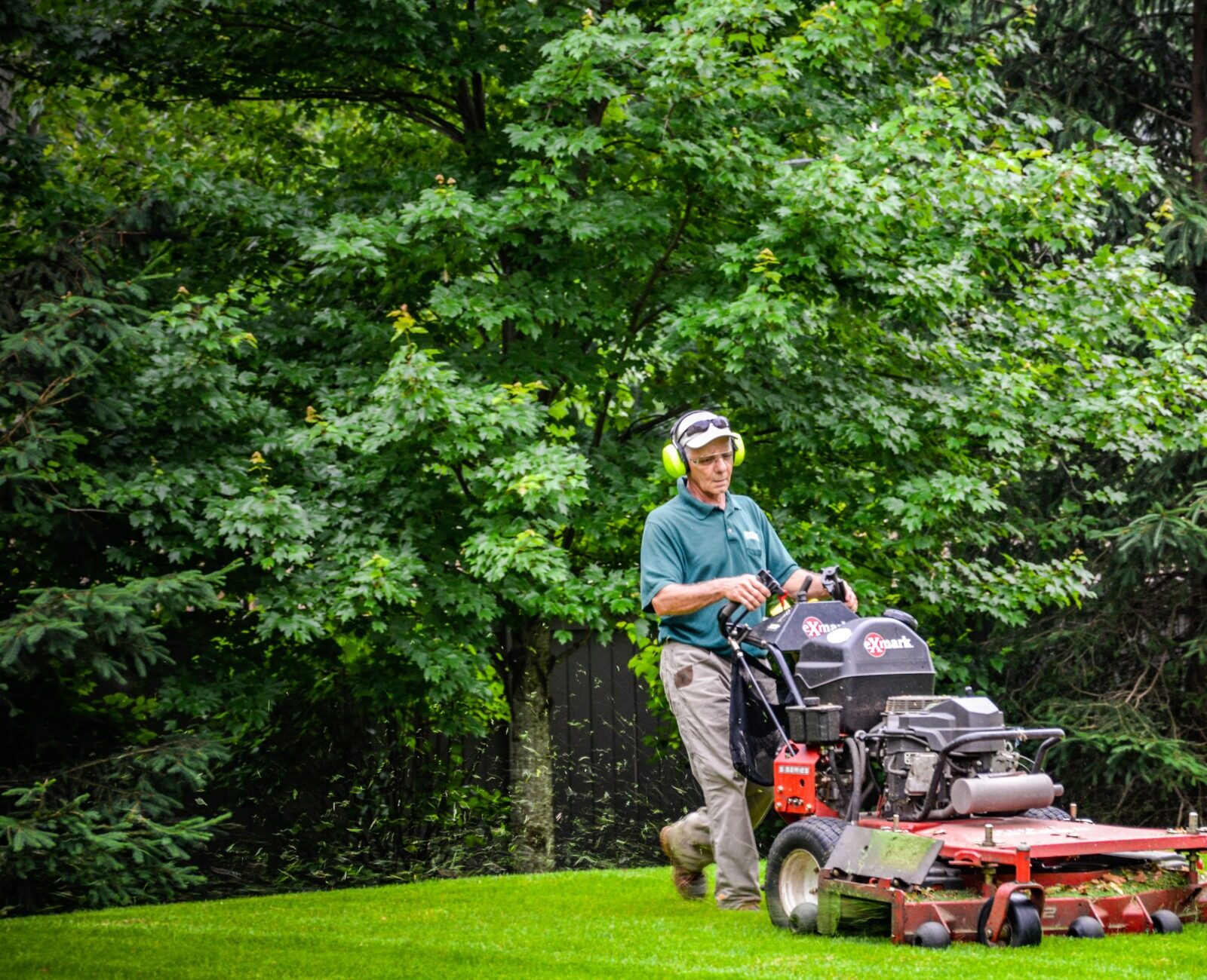A person mows a lush green lawn using a red commercial riding lawn mower, with dense trees in the background during daylight.