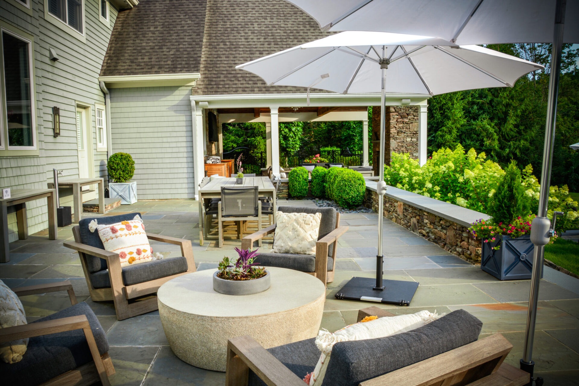 An elegant outdoor patio with comfortable seating, a dining area under an umbrella, lush greenery, and a serene atmosphere adjacent to a house.