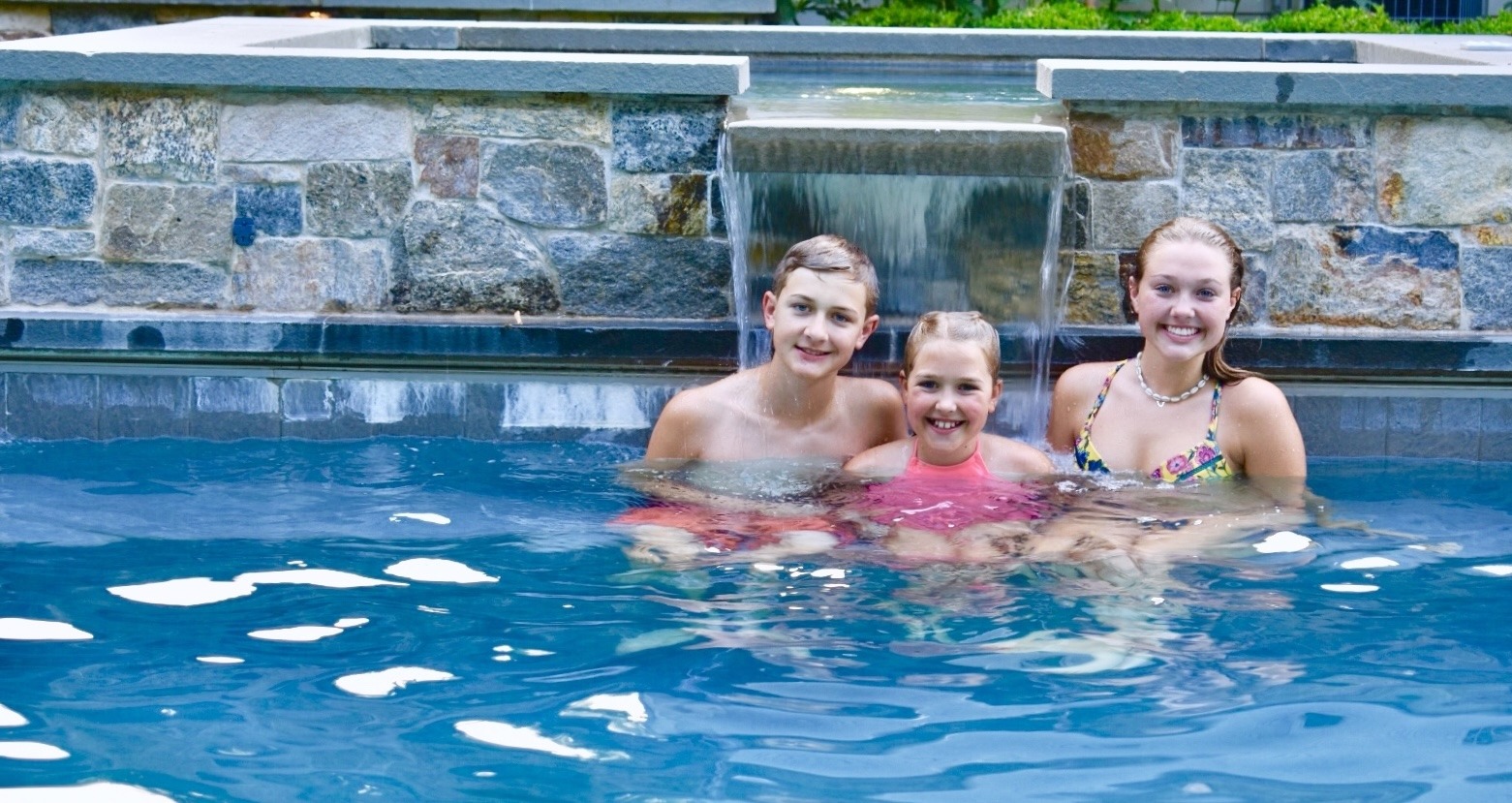 Three smiling young individuals are in a swimming pool with a stone waterfall feature behind them. They appear to be enjoying a leisurely swim.