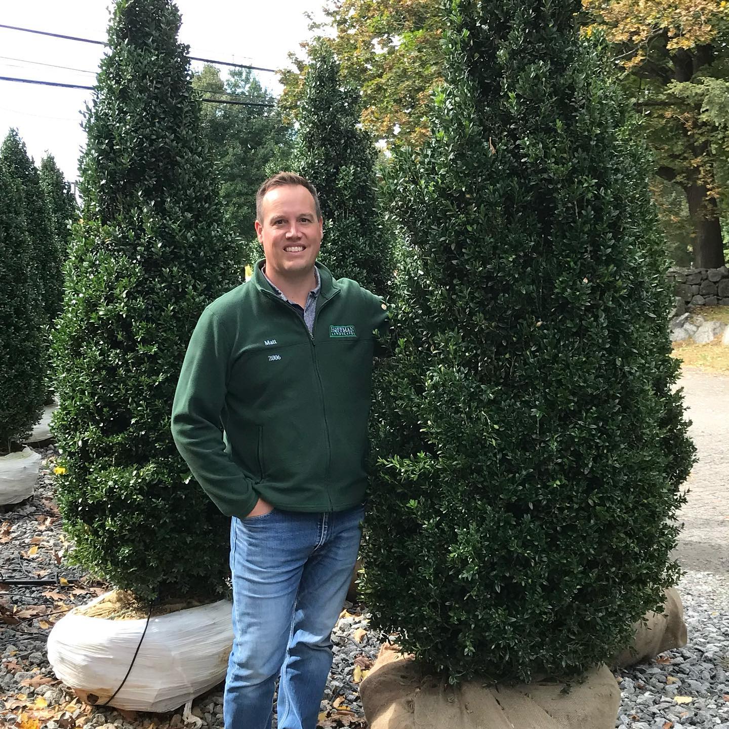 A smiling person in a green fleece jacket stands among tall, conical shrubs wrapped at the base, outdoors on a clear day.