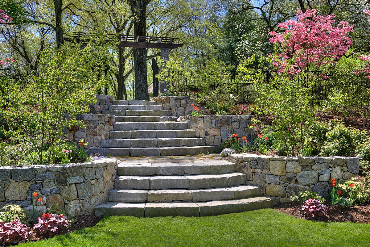 Stone staircase with retaining walls amidst lush garden, blooming pink and white flowers, greenery under a clear sky. Visually inviting outdoor landscape with architectural elements.