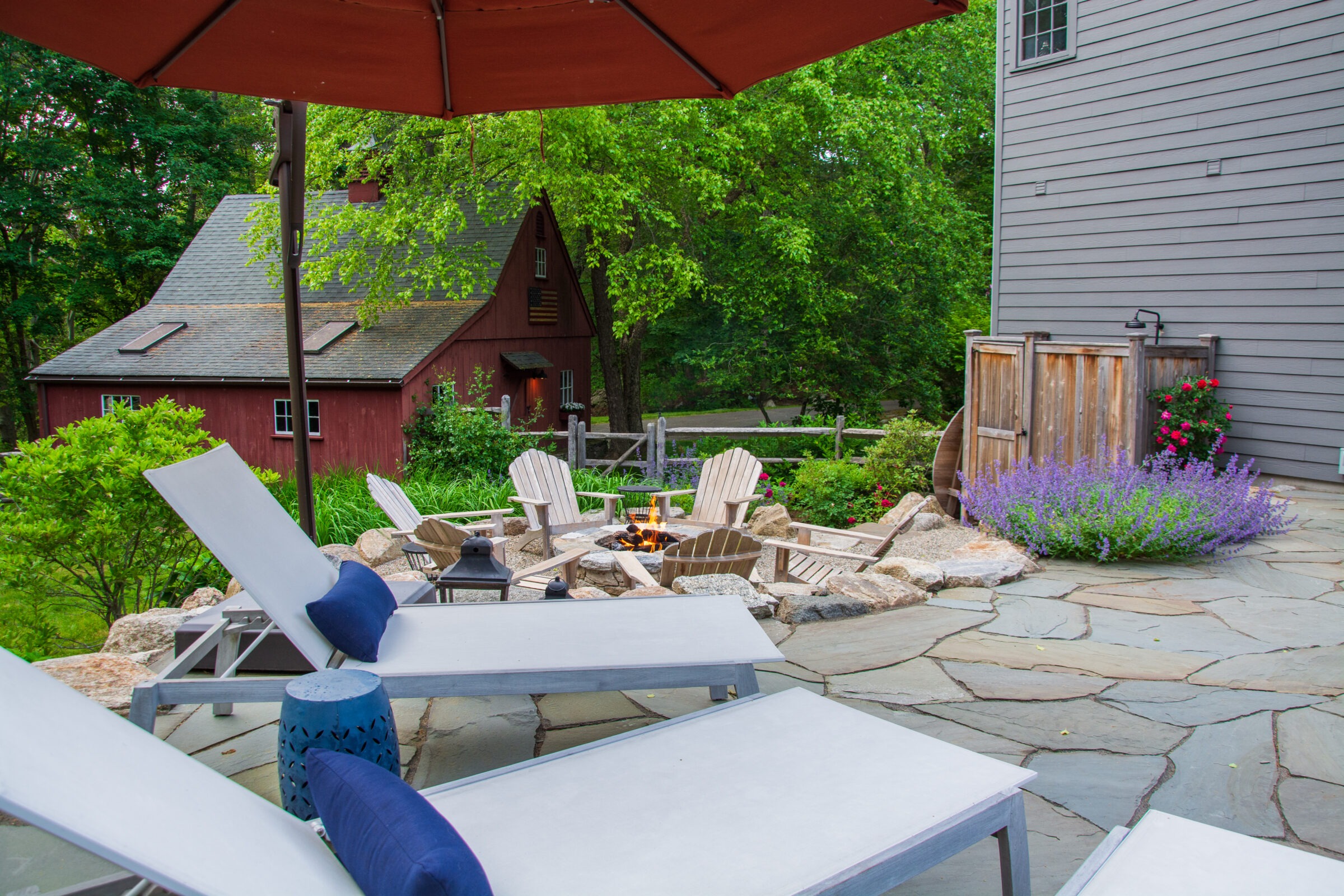 A cozy outdoor patio with lounge chairs, a fire pit, colorful flowers, and a rustic red barn in the background surrounded by lush greenery.