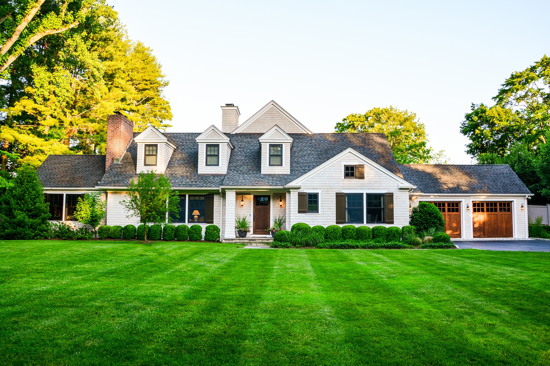 This is an image of a two-story suburban house with a well-manicured lawn, white siding, grey shingles, and an attached garage in a peaceful setting.