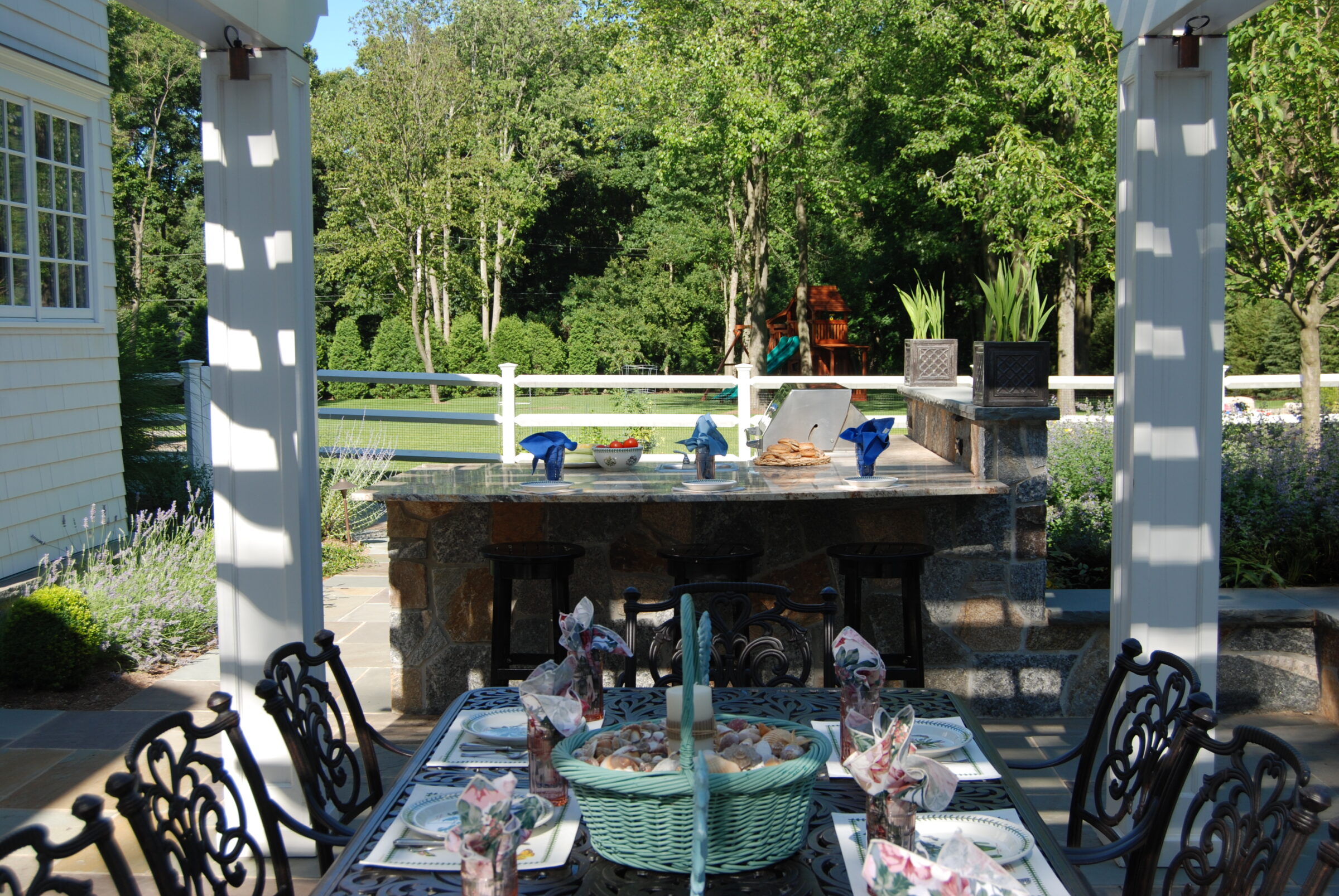 An outdoor dining area with a stone bar, metal chairs, and a set table under a pergola. Greenery surrounds this tranquil patio setting.