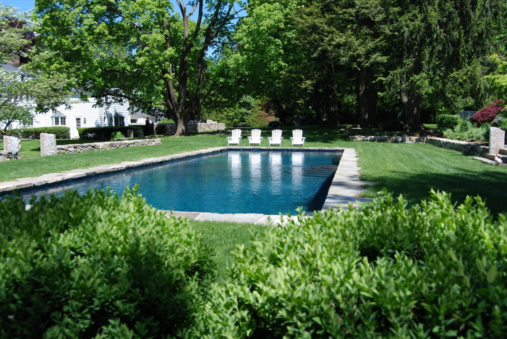 An outdoor swimming pool with clear blue water, surrounded by lush greenery, white chairs lined up, and a traditional white house in the background.