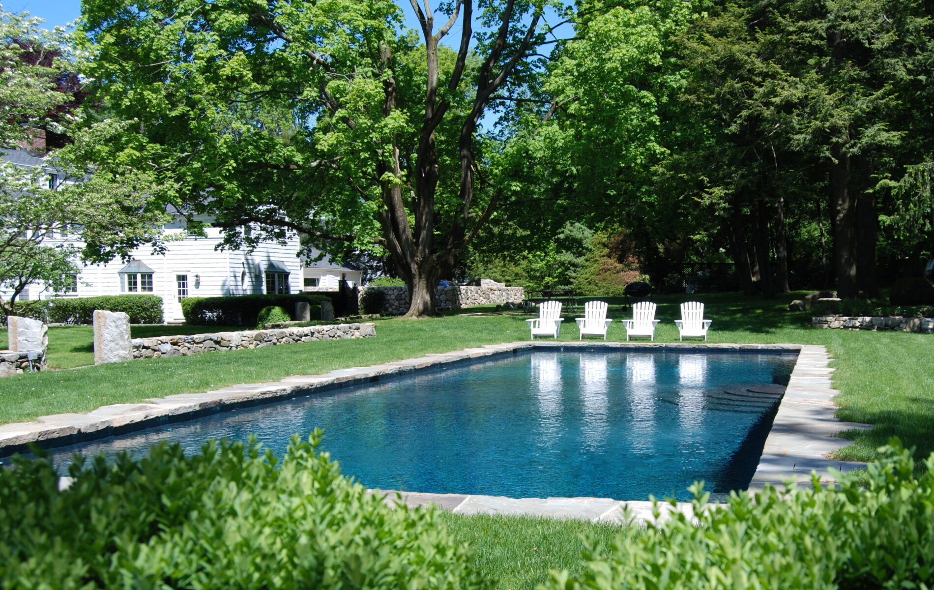 An outdoor swimming pool with clear blue water, surrounded by lush greenery, white chairs lined up, and a traditional white house in the background.
