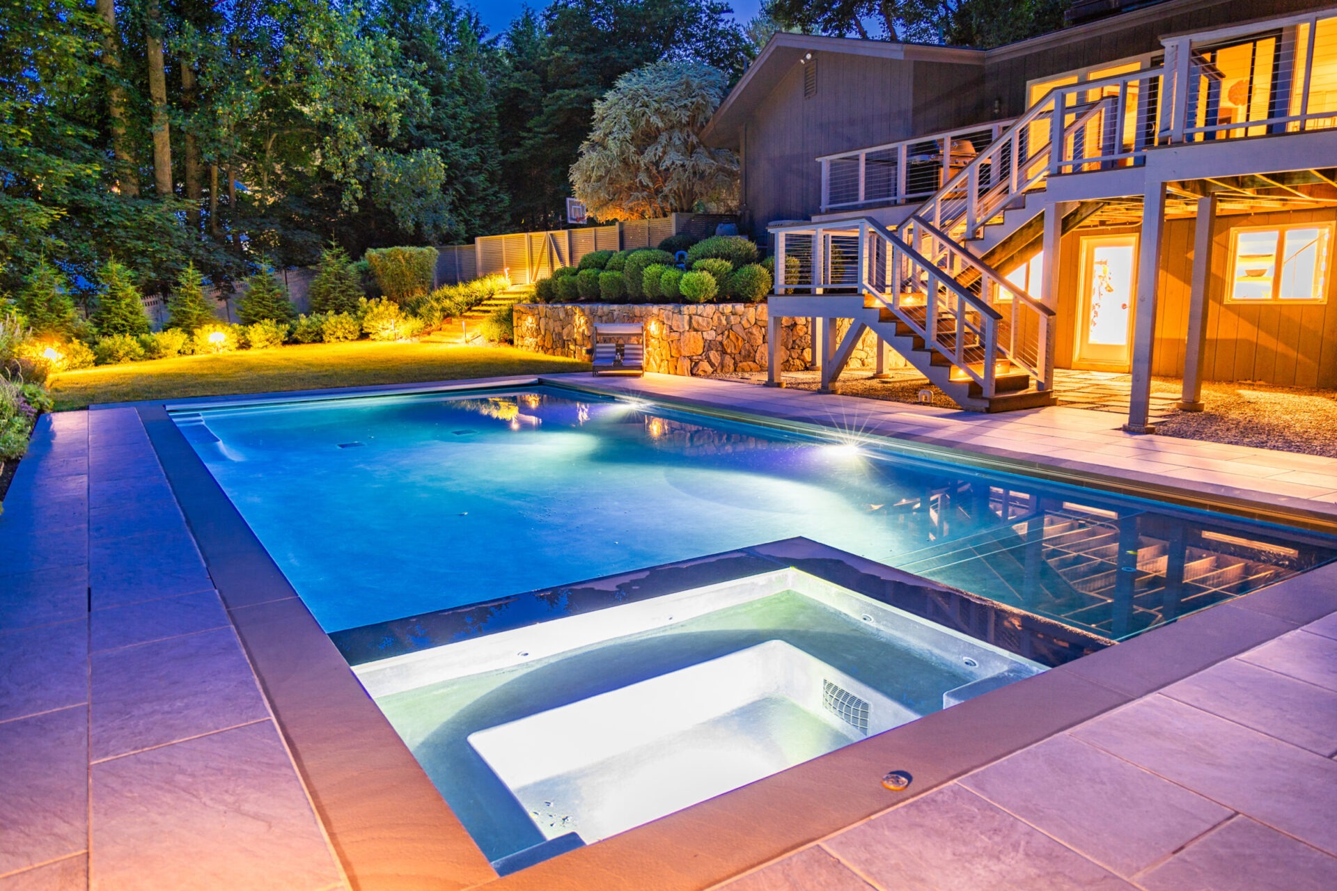 Modern backyard pool and hot tub illuminated at dusk, surrounded by lush landscaping, uplighting, and a two-level deck attached to a contemporary home.
