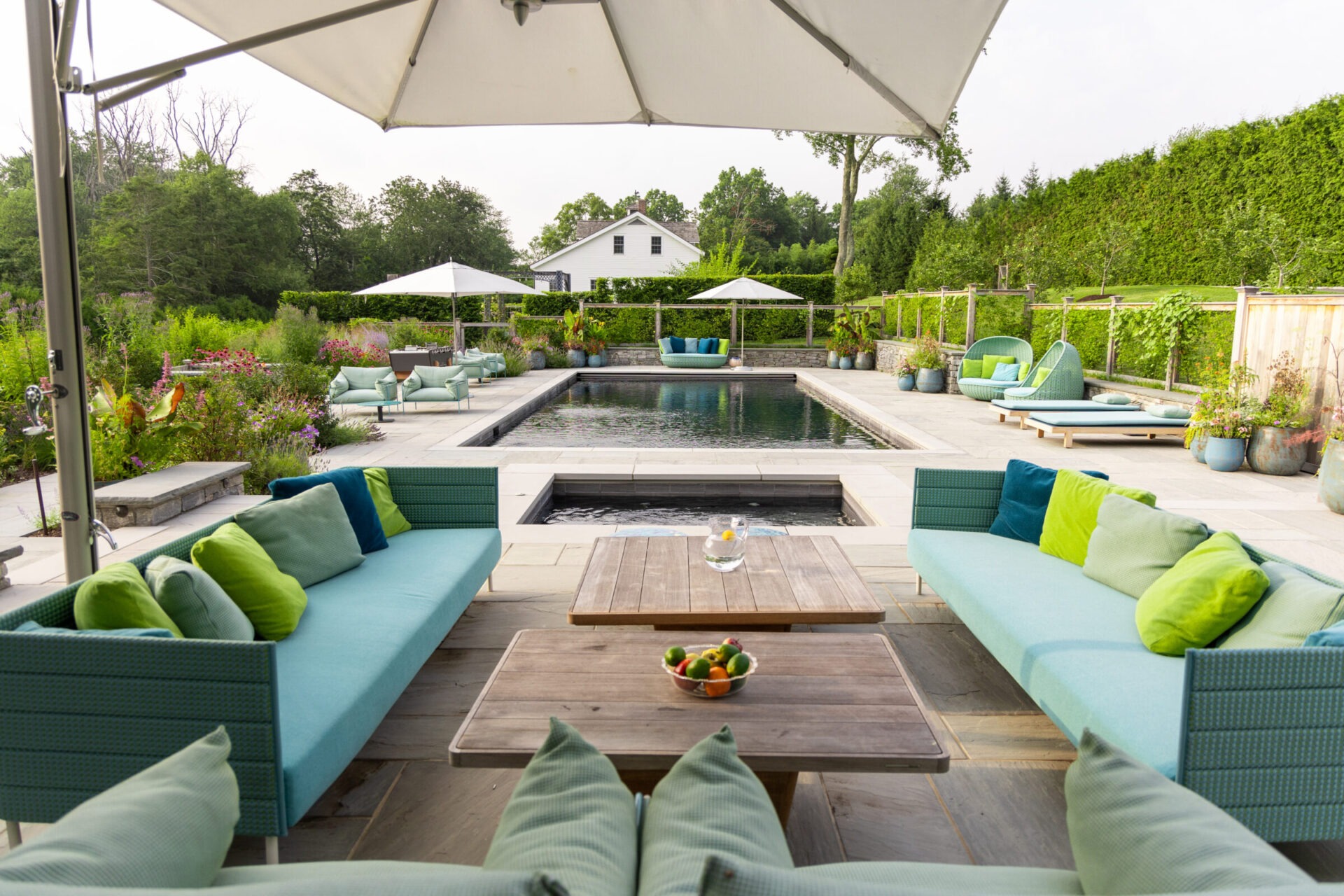 Luxury backyard pool area with vibrant landscaping, modern teal and green outdoor furniture, a hot tub in the foreground, and a rectangular pool flanked by lounge chairs and large white umbrellas, all set against a backdrop of trees and hedges.