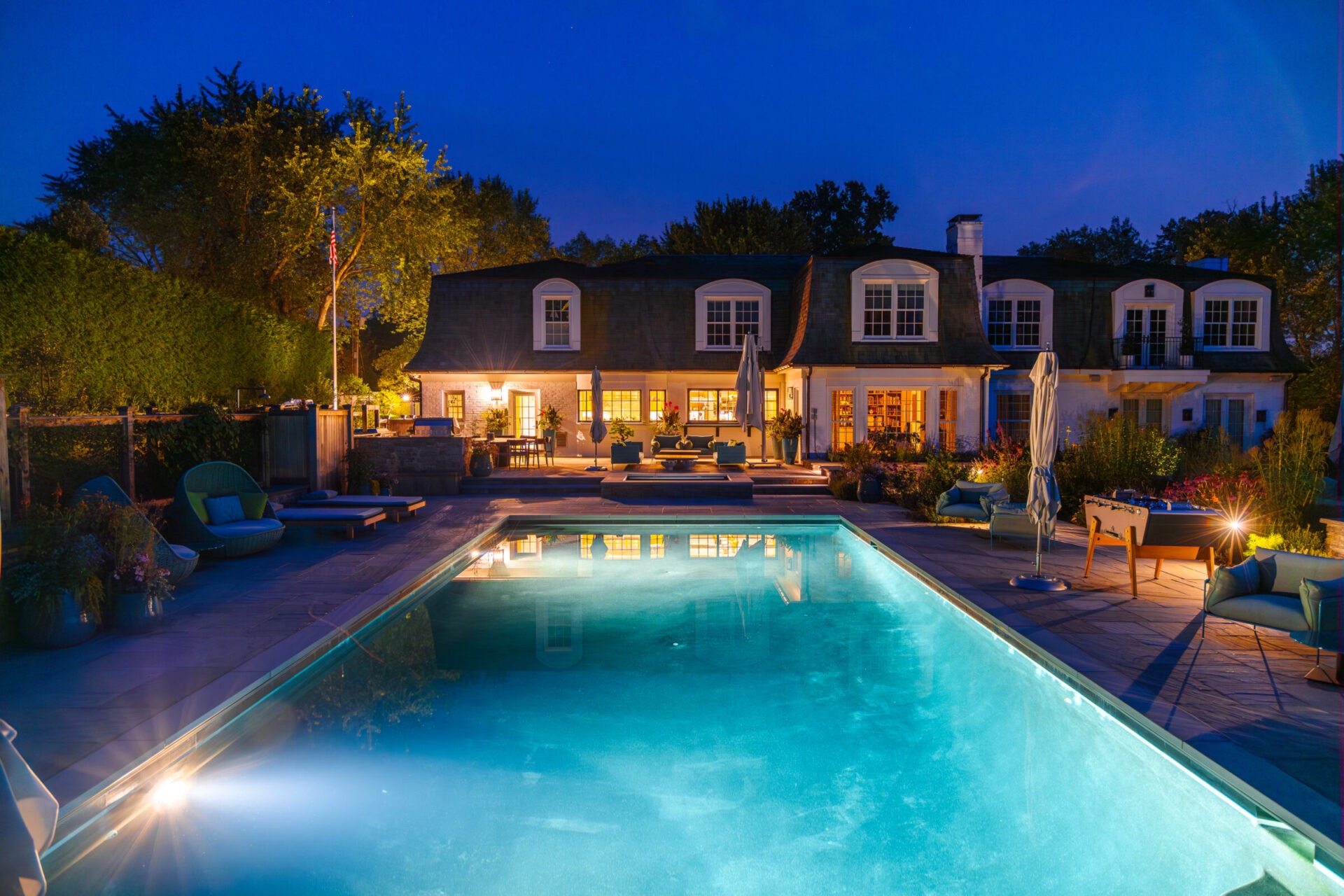 Elegant backyard with a glowing swimming pool at night, surrounded by cozy lounge seating, landscaping lights, and a large two-story traditional-style home with dormer windows and warm interior lighting.