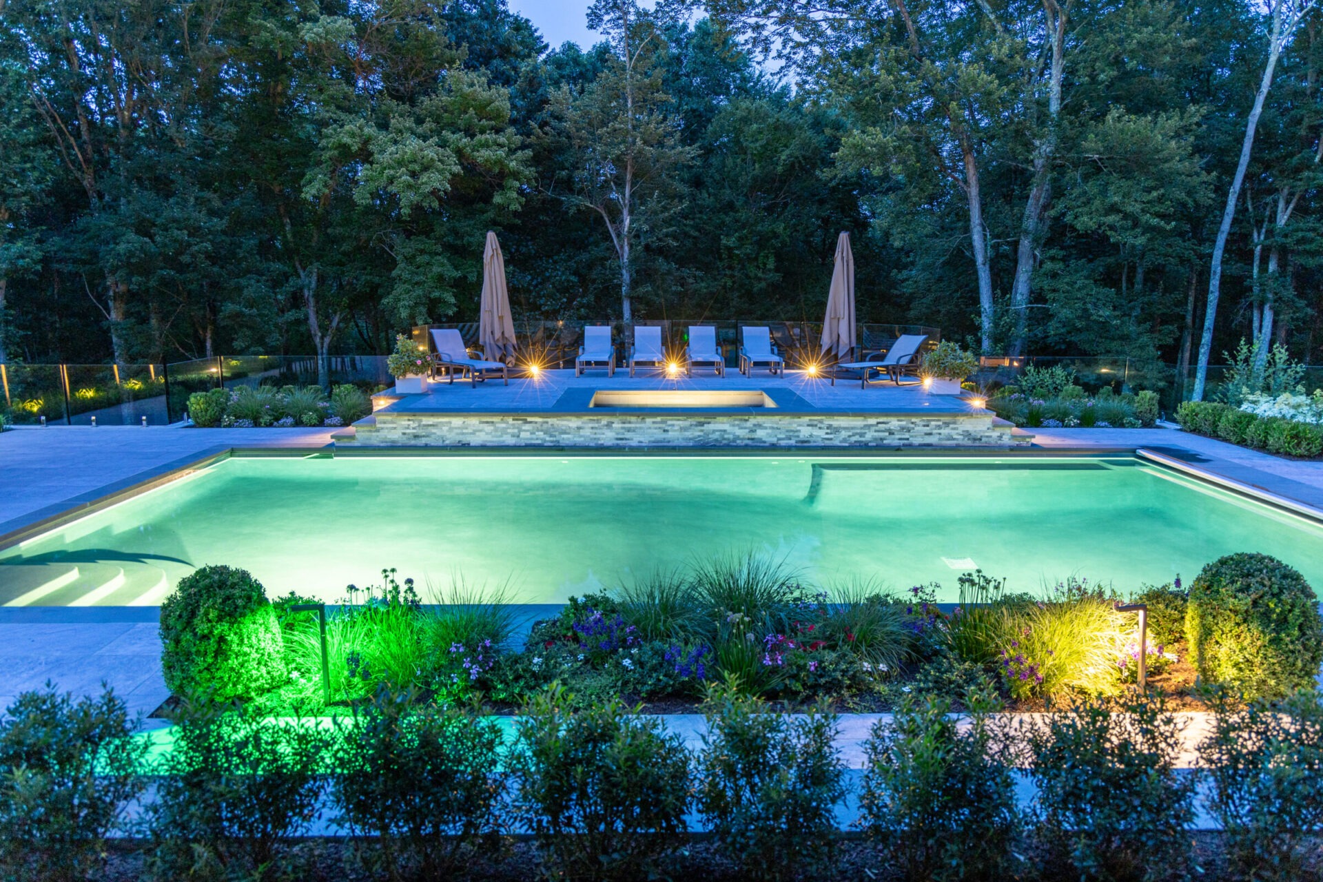 Luxurious backyard with a glowing in-ground pool, vibrant landscape lighting, manicured gardens, and a modern poolside lounge area surrounded by trees.