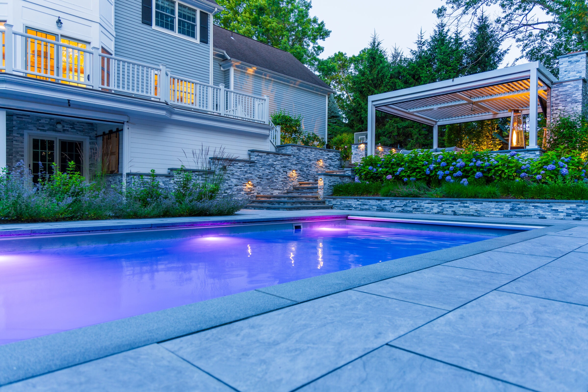 Modern backyard with a glowing purple-lit swimming pool, stone steps, lush landscaping with hydrangeas, and a covered outdoor lounge area with warm lighting and a fireplace.