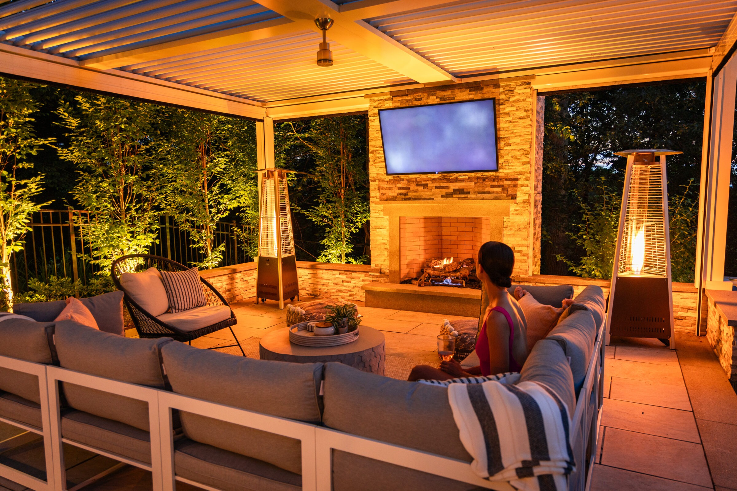 Woman relaxing with a drink in a cozy covered outdoor lounge featuring a sectional sofa, fire features, a mounted TV above a brick fireplace, and ambient lighting highlighting the surrounding trees.