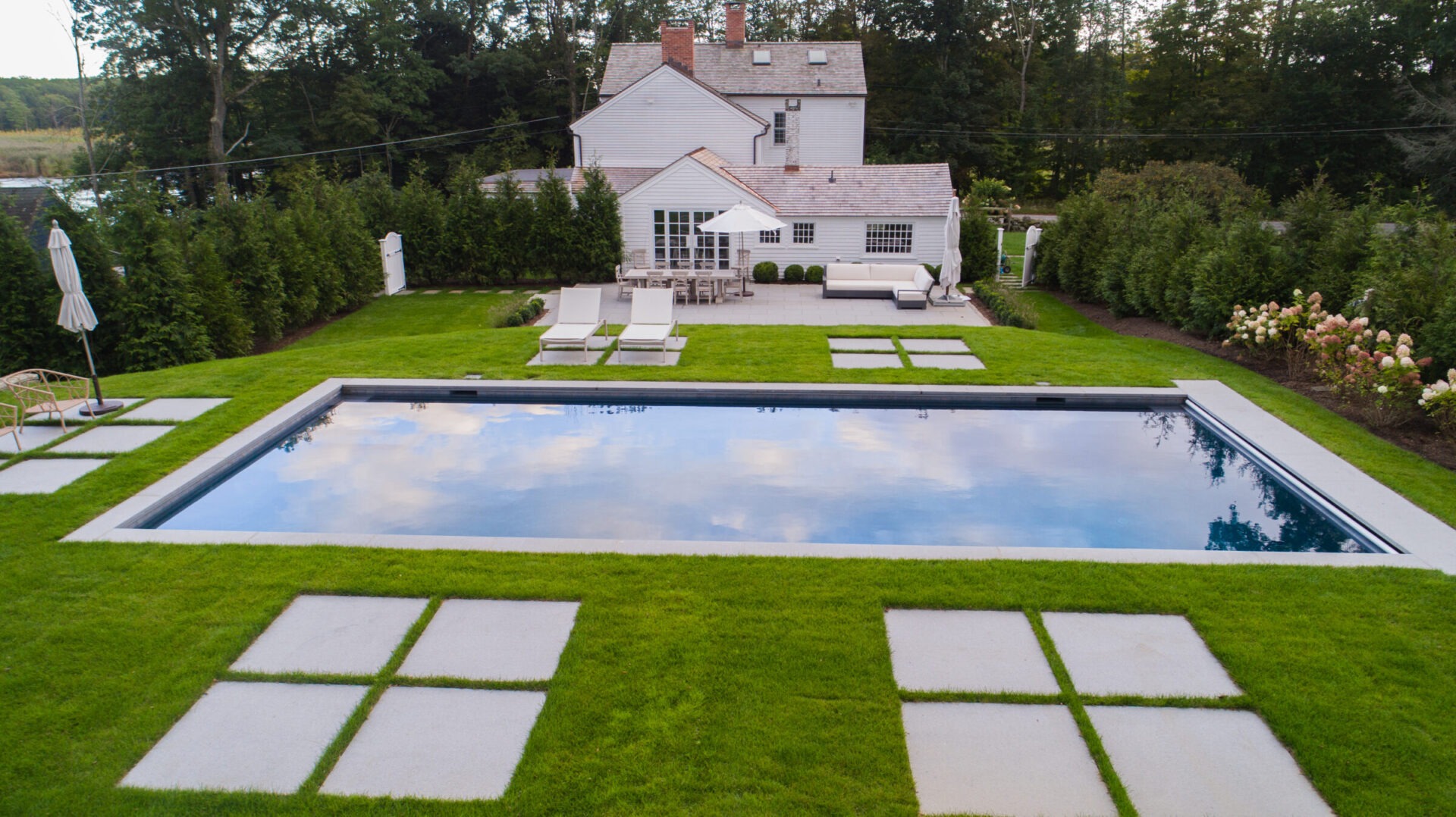 Modern backyard with a rectangular inground pool surrounded by lush green grass, concrete pavers, lounge chairs, and a patio area with outdoor seating and a white umbrella in front of a white house.