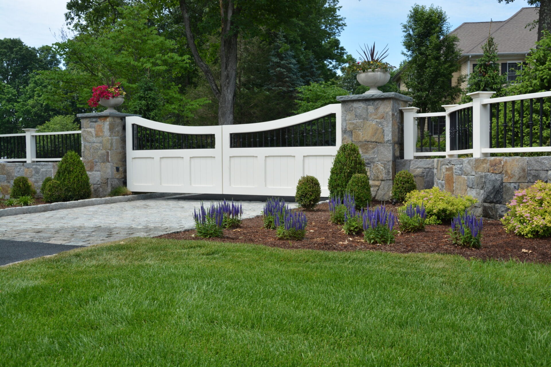 Elegant white driveway gate with black iron accents, flanked by stone columns topped with decorative planters filled with colorful flowers, set against a backdrop of manicured landscaping with purple flowering plants and trimmed shrubs.