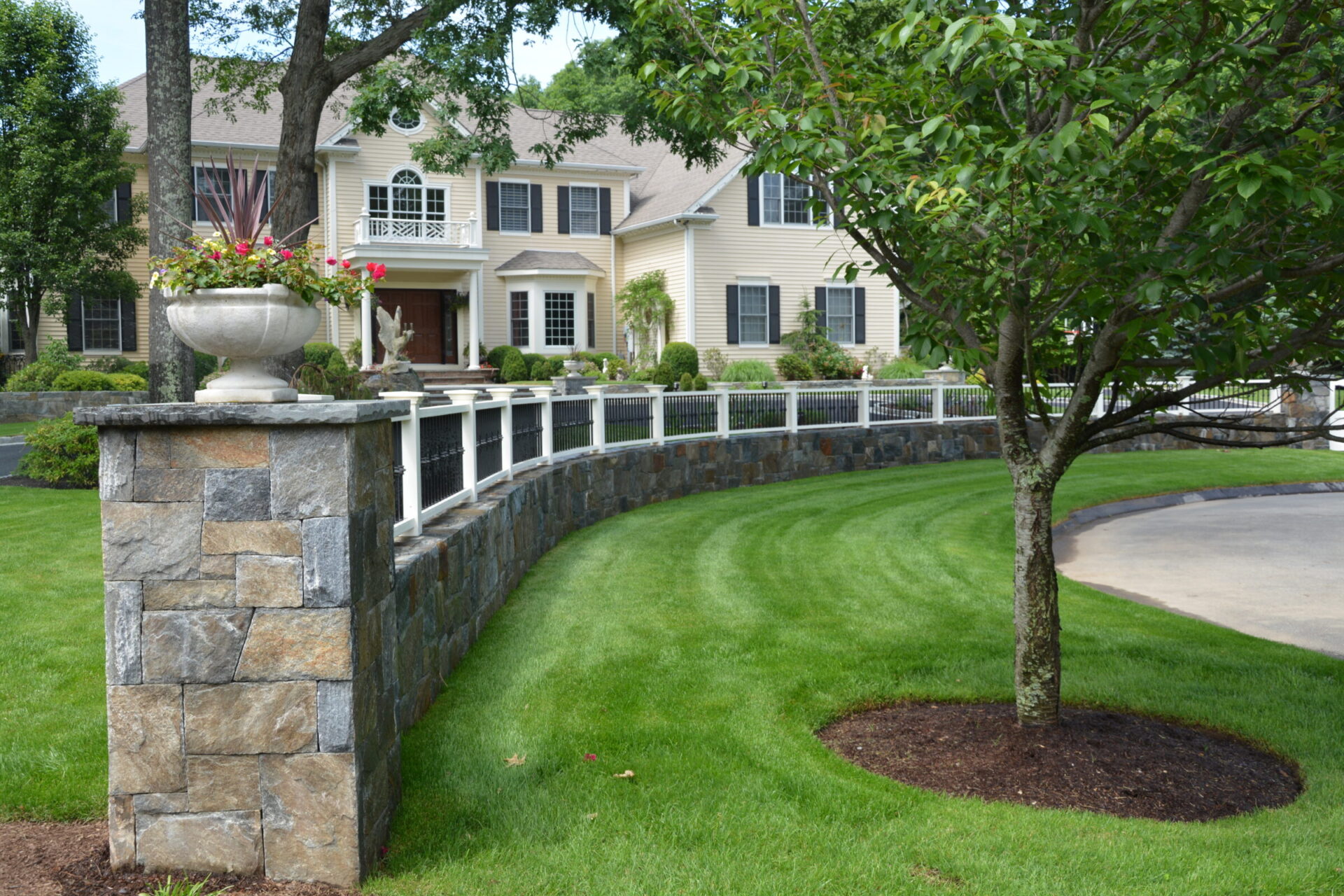 Beautifully landscaped front yard of a large beige colonial-style home with black shutters, featuring a curved stone retaining wall topped with a white and black iron fence, lush green lawn, and decorative planter with pink flowers.