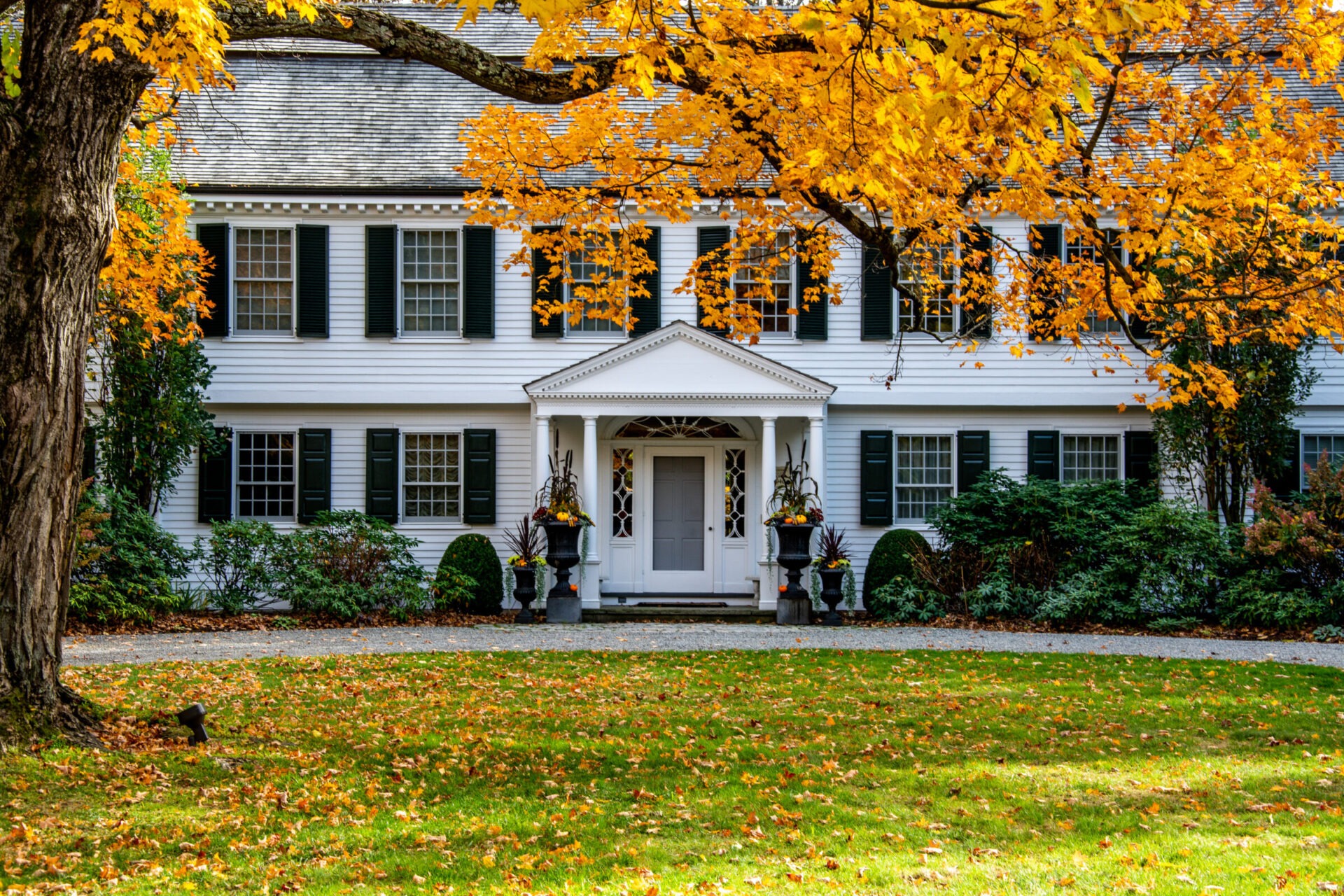 Classic white colonial-style home with black shutters and a symmetrical facade, framed by vibrant autumn foliage and a lawn scattered with fallen leaves, featuring a central portico entrance flanked by tall decorative planters.