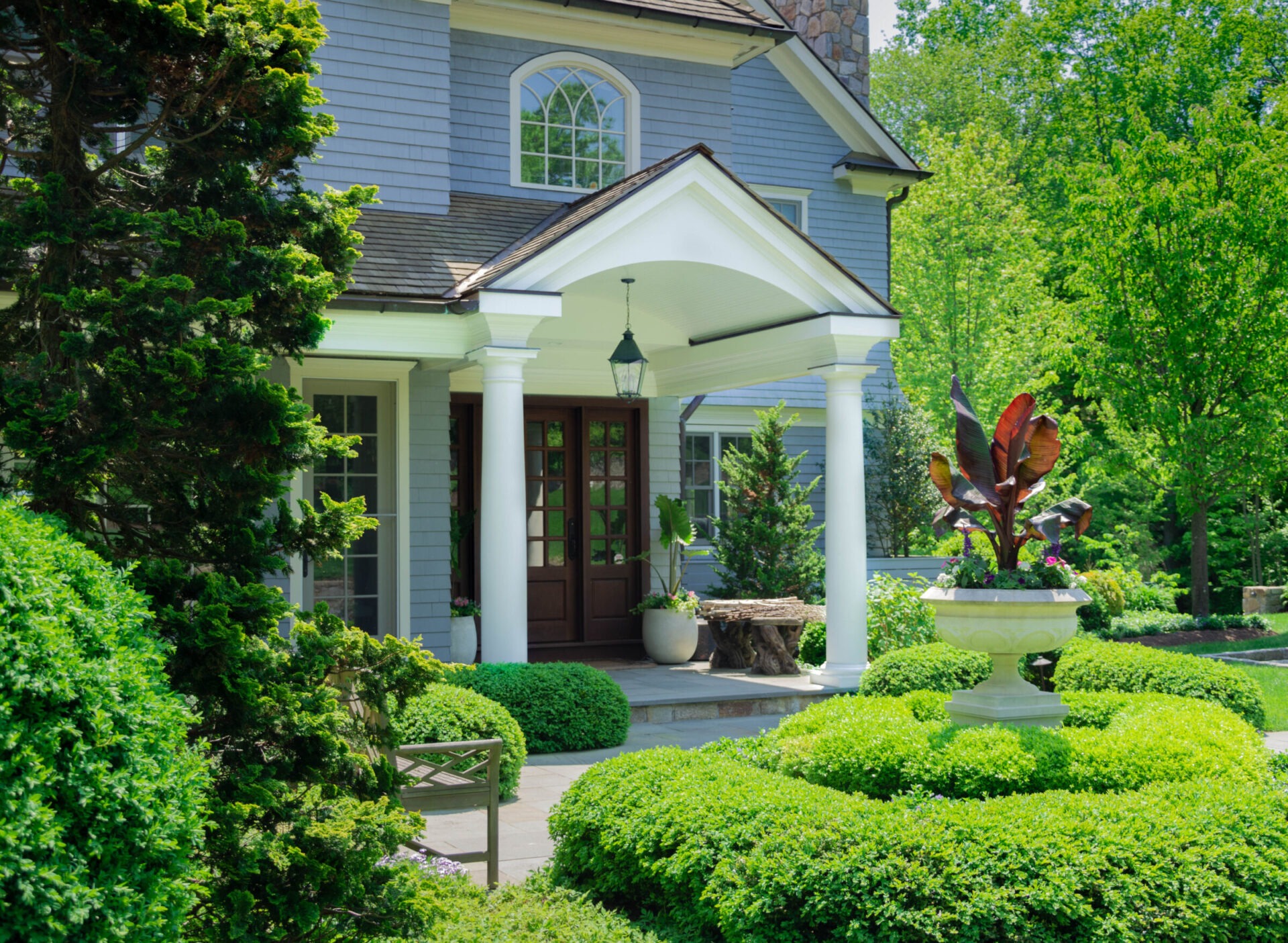 Elegant home entrance with white columns, arched portico, and manicured landscaping featuring lush green shrubs and a large decorative planter with tropical foliage as a focal point.