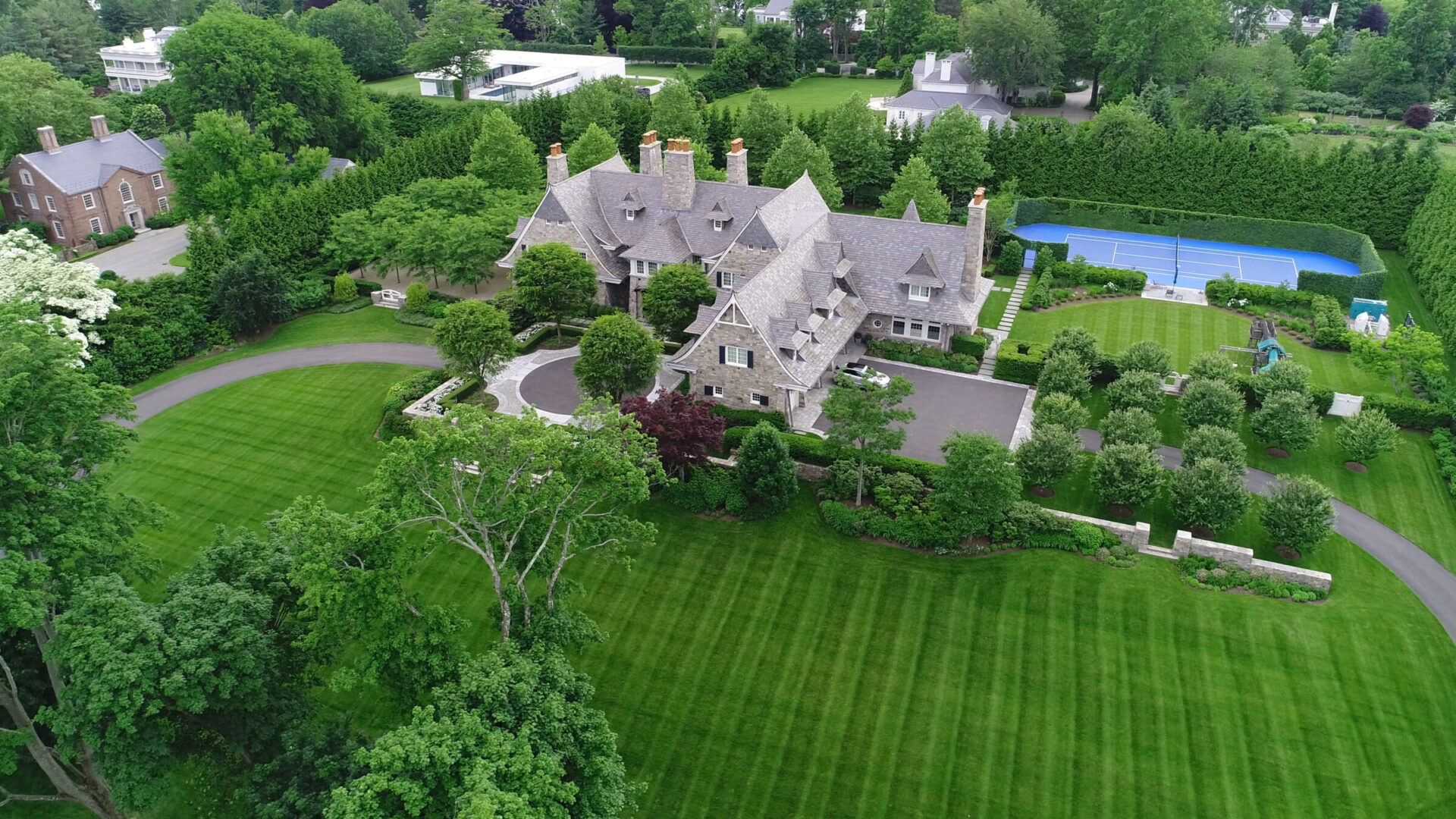 Aerial view of a sprawling estate with a stone mansion, circular driveway, manicured lawns, mature trees, a tennis court, and a private playground, surrounded by hedges and other large homes.